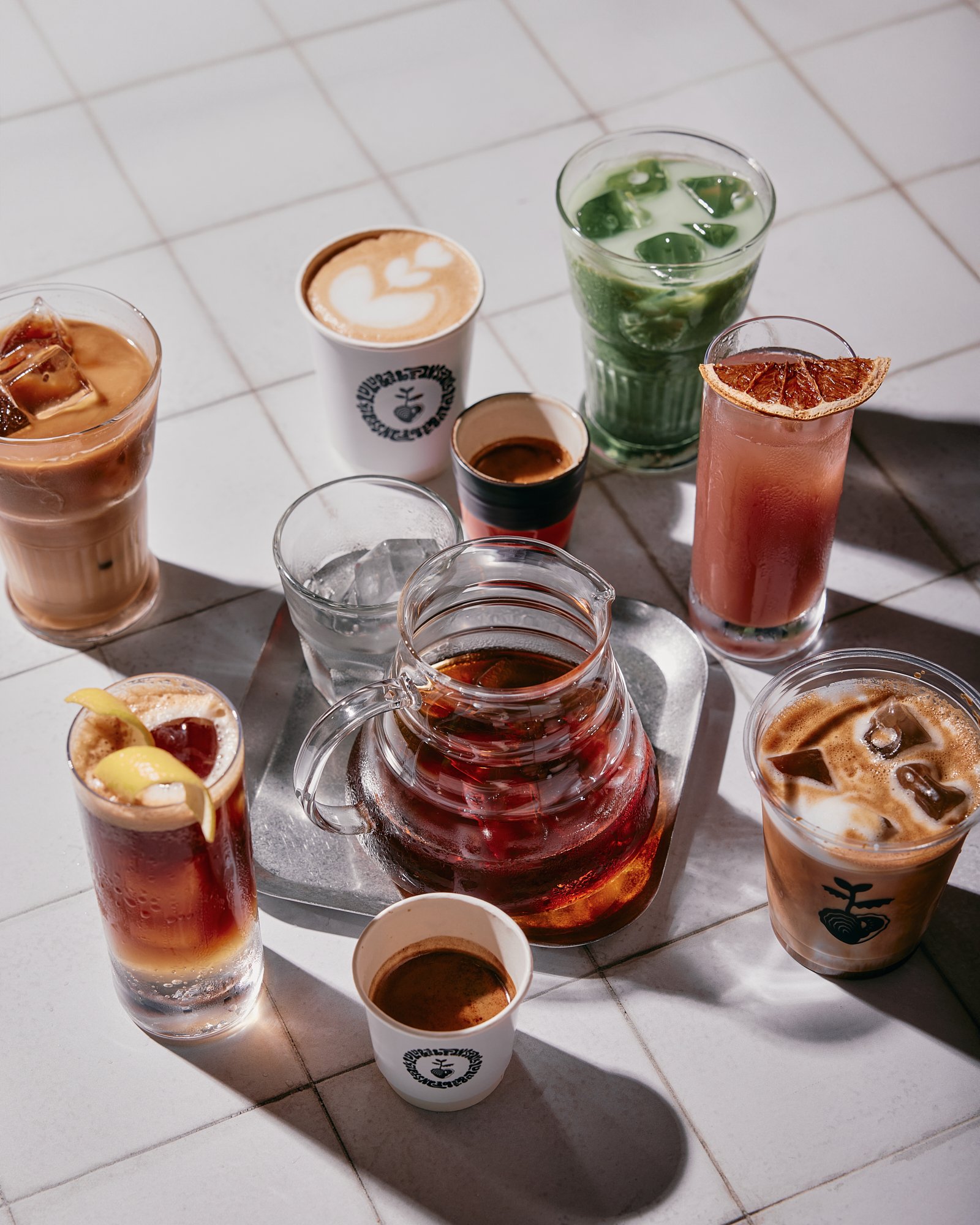 A variety of coffee drinks and beverages on a white tiled surface, including iced coffee, espresso, latte, tea, and colorful drinks with garnishes.