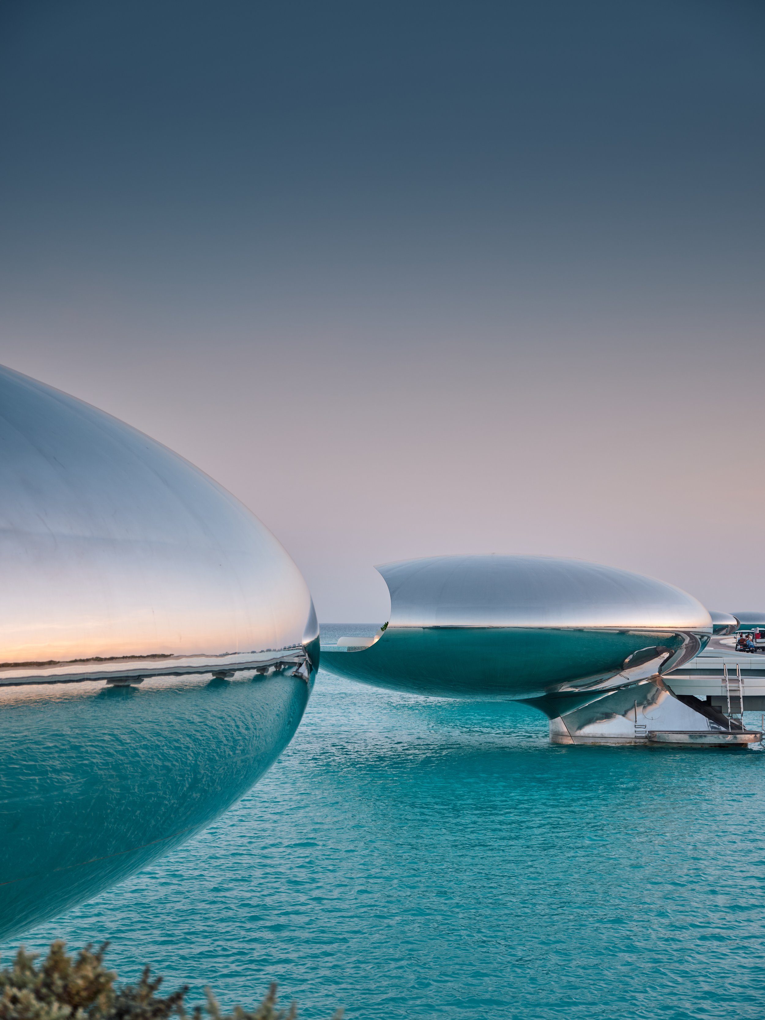 Two modern, metallic sculptures resembling futuristic ships or domes are positioned on the water at a marina, with the sky transitioning from sunset to dusk in the background.
