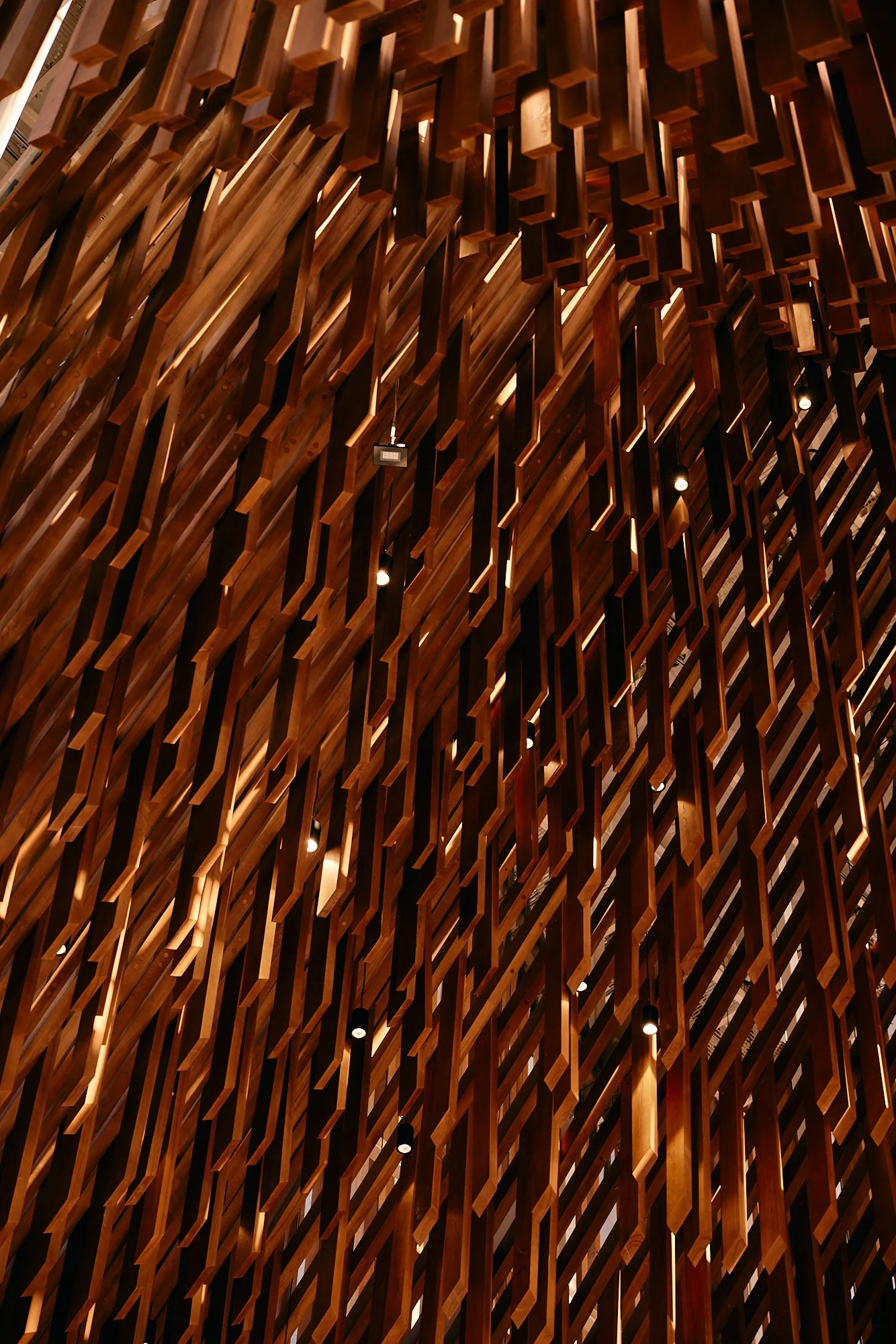 Interior decorative installation made of numerous vertical wooden slats arranged in a flowing, wave-like pattern with small spotlights focused on the wood.