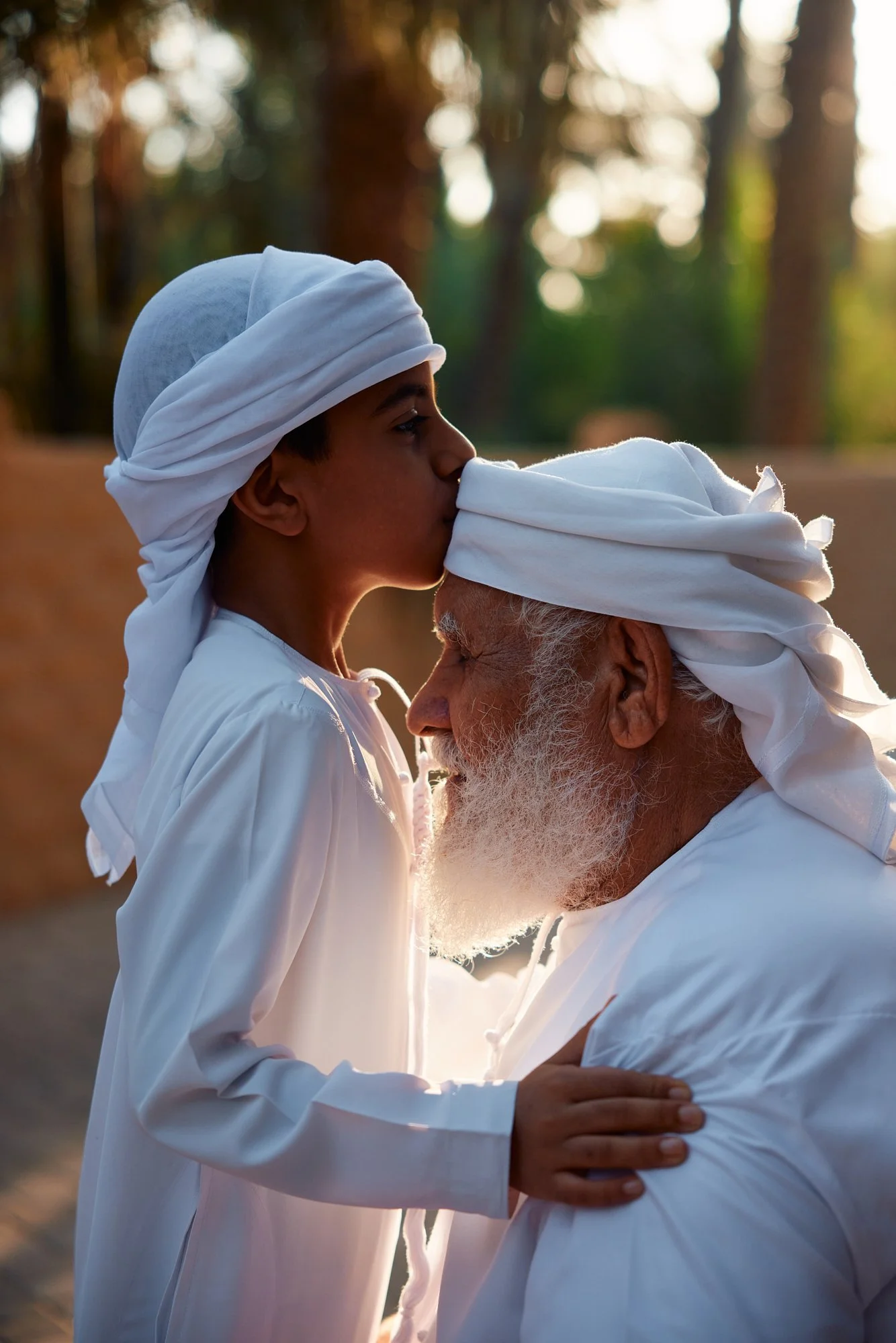 A young boy giving a kiss on the forehead to an elderly man, both dressed in traditional white clothing and head coverings, outdoors with trees and sunlight in the background.