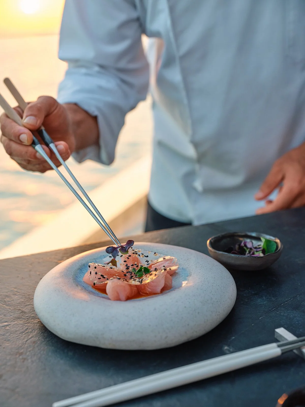 Person in a light blue shirt using tweezers to place edible flowers on a plated dish of sashimi on a stone plate during sunset by the water.