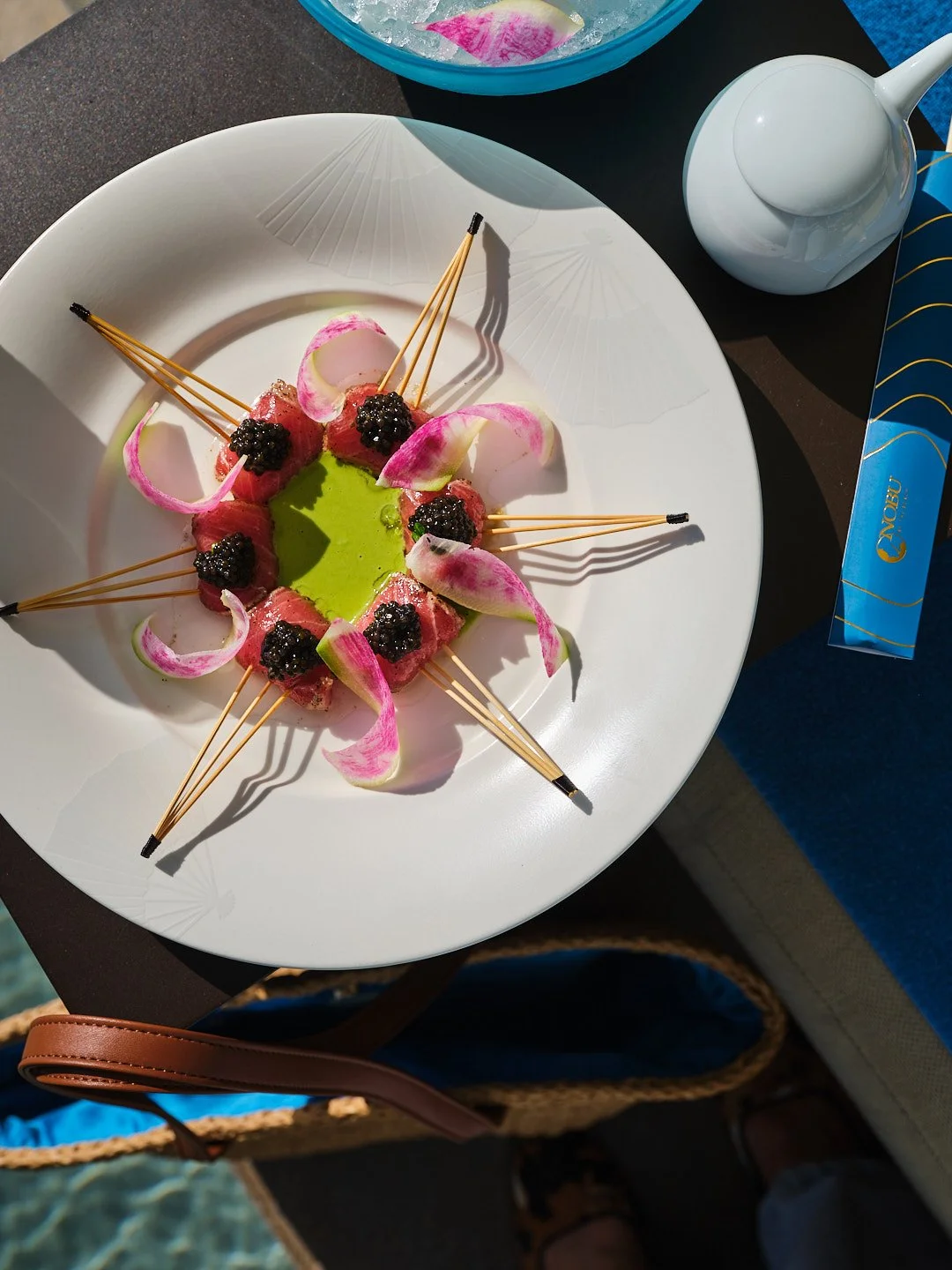 A white plate with a decorative pattern, holding a circular arrangement of pink and green food garnished with blackberries and pink edible flower petals, with skewers sticking out from the toppings. There is a teapot and a blue bowl with ice and a half lemon in the background.