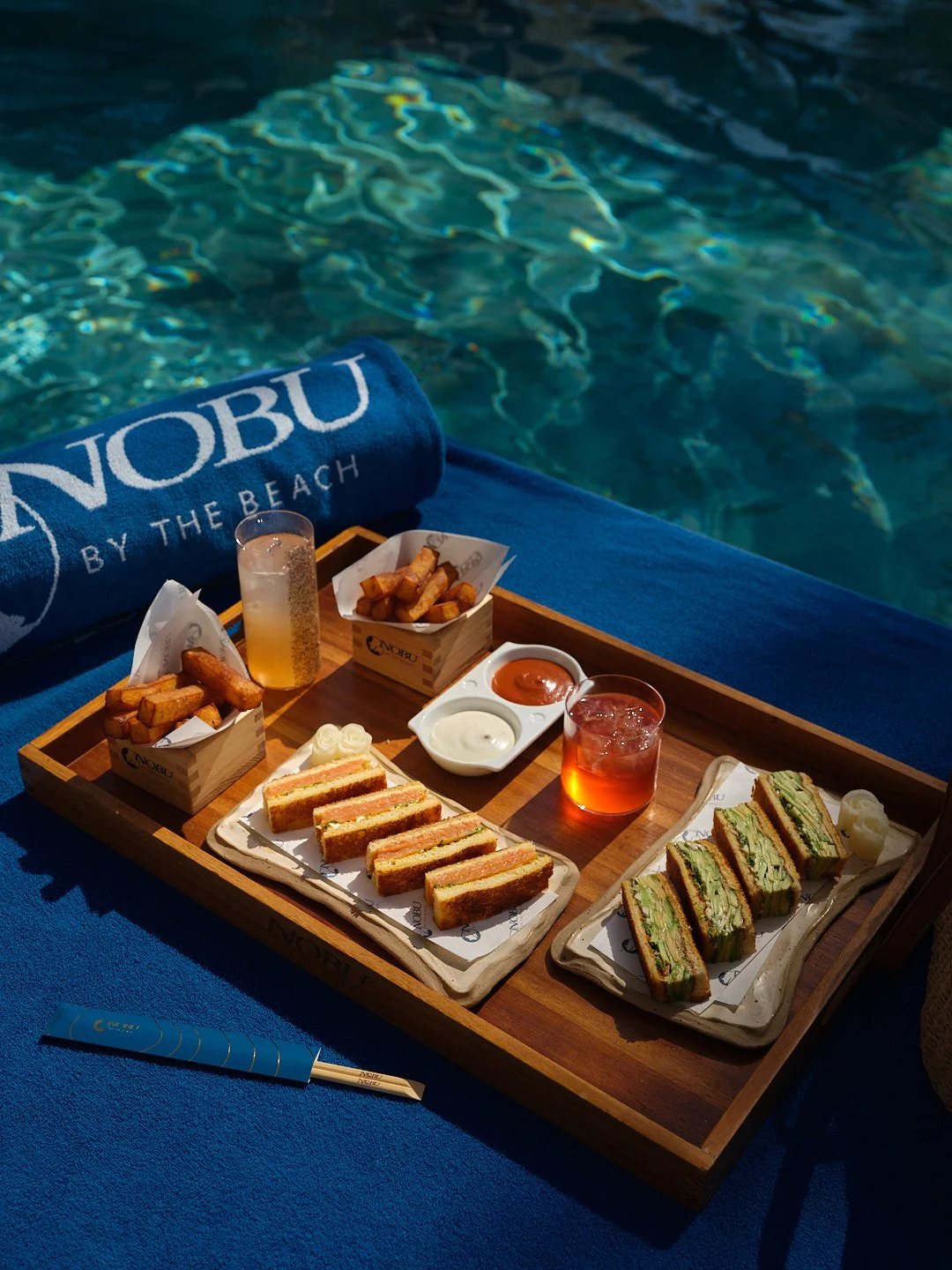 A wooden tray with sandwiches, fries, sauces, a drink, and a towel with the words 'NOBU BY THE BEACH' lies on a blue surface next to a pool.