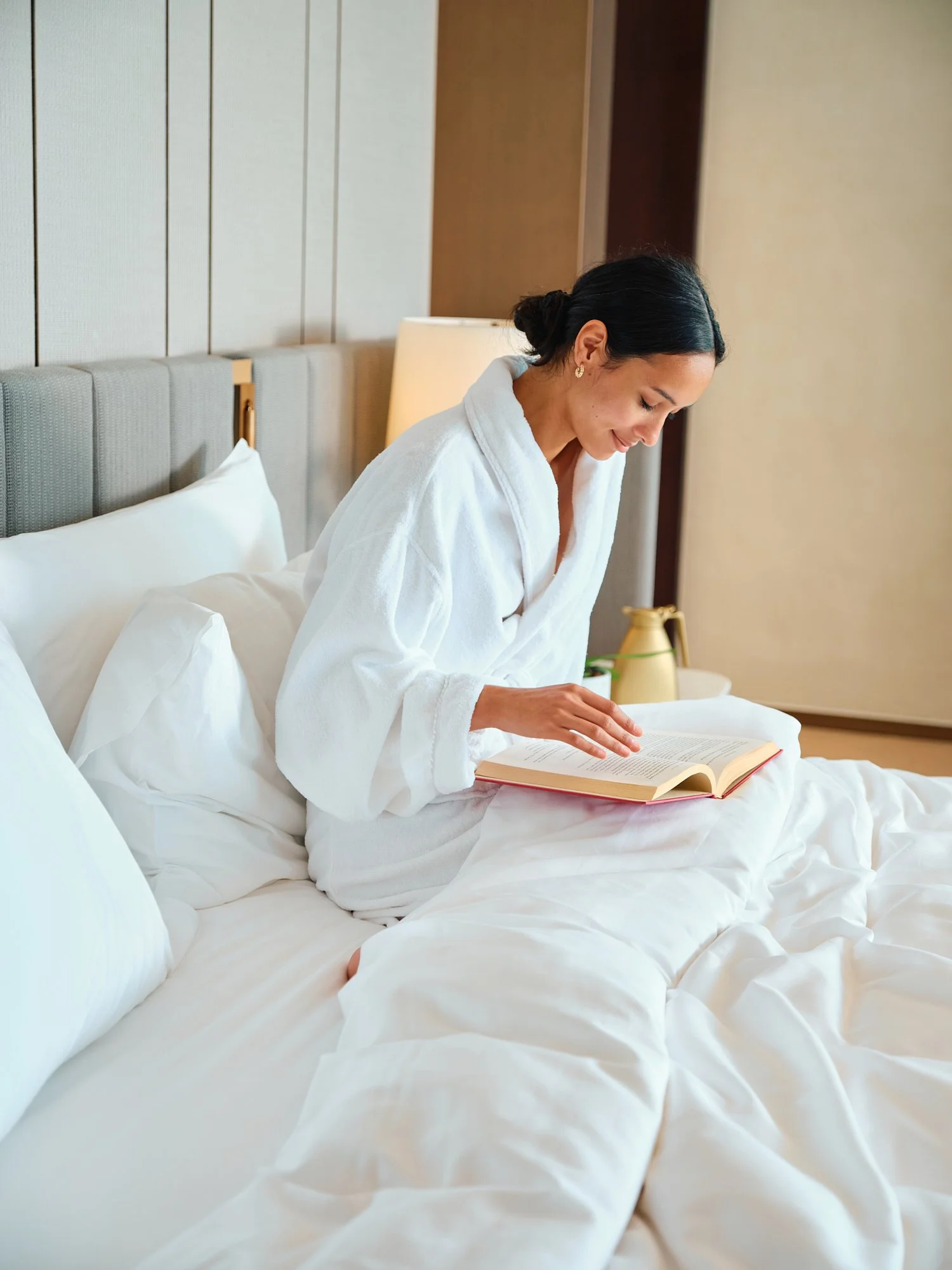 A woman in a white bathrobe sitting on a bed, reading a book.