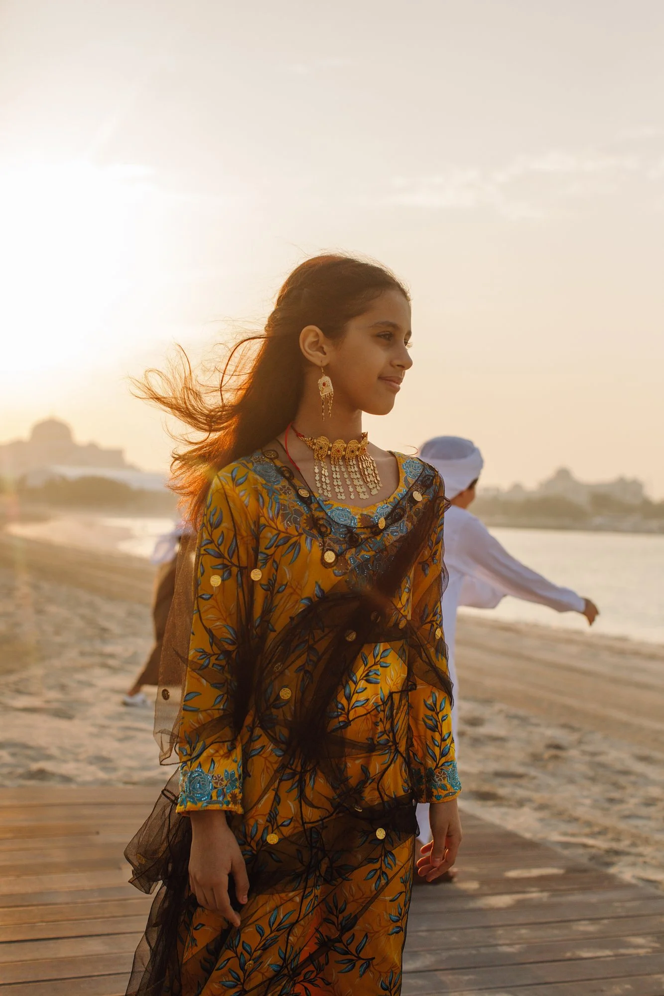 A young woman in a yellow embroidered dress with blue and black accents, wearing gold jewelry, standing on a beach during sunset with wind blowing her hair.