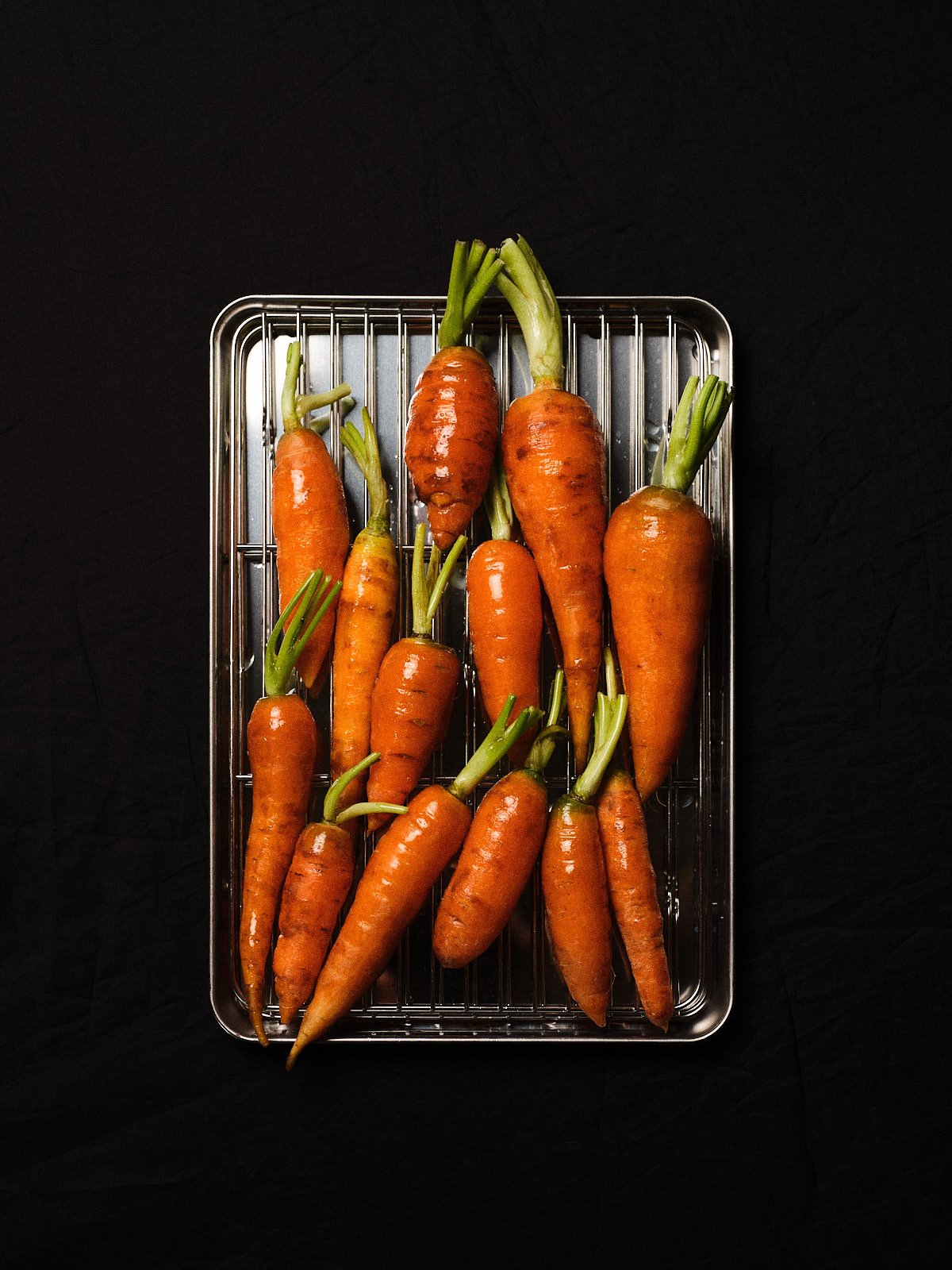 Fresh orange carrots with green tops on a metal wire rack against a black background.