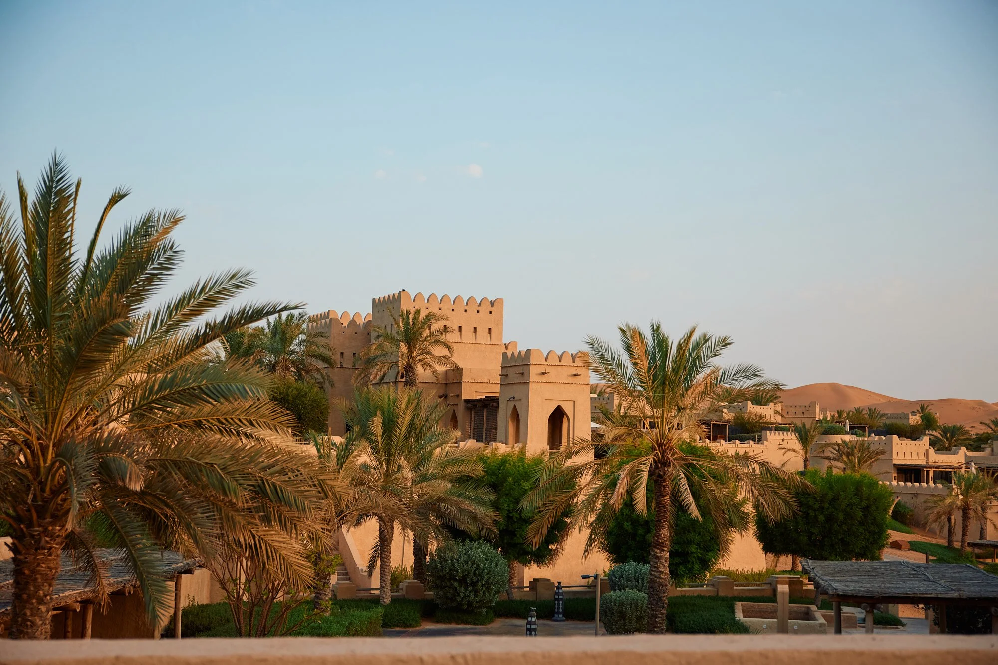 Desert fortress surrounded by palm trees and desert dunes in the background at sunset.