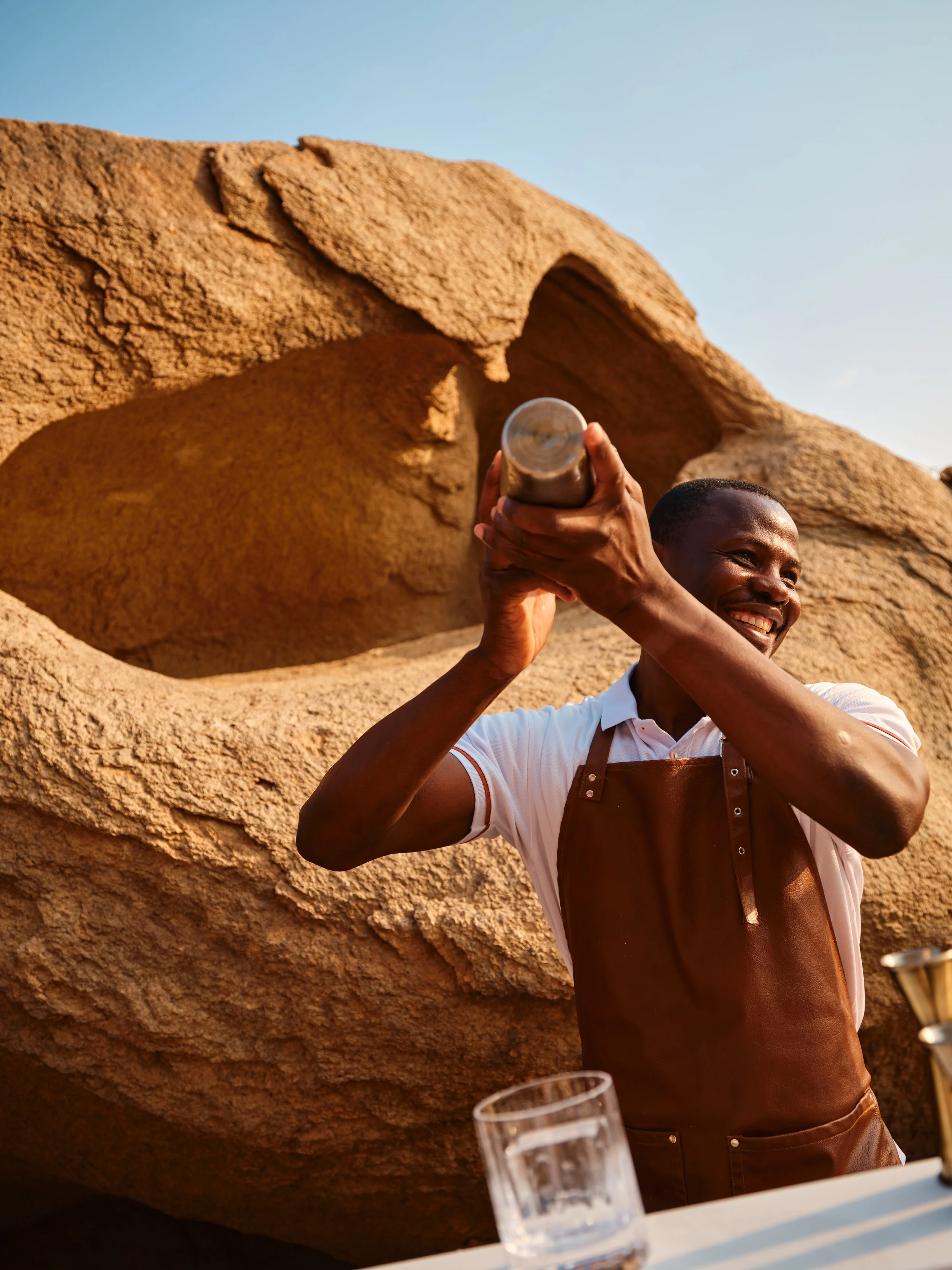 A smiling man wearing a white shirt and brown apron shakes a metal cocktail shaker outdoors at sunset, with large rocks and blue sky in the background.