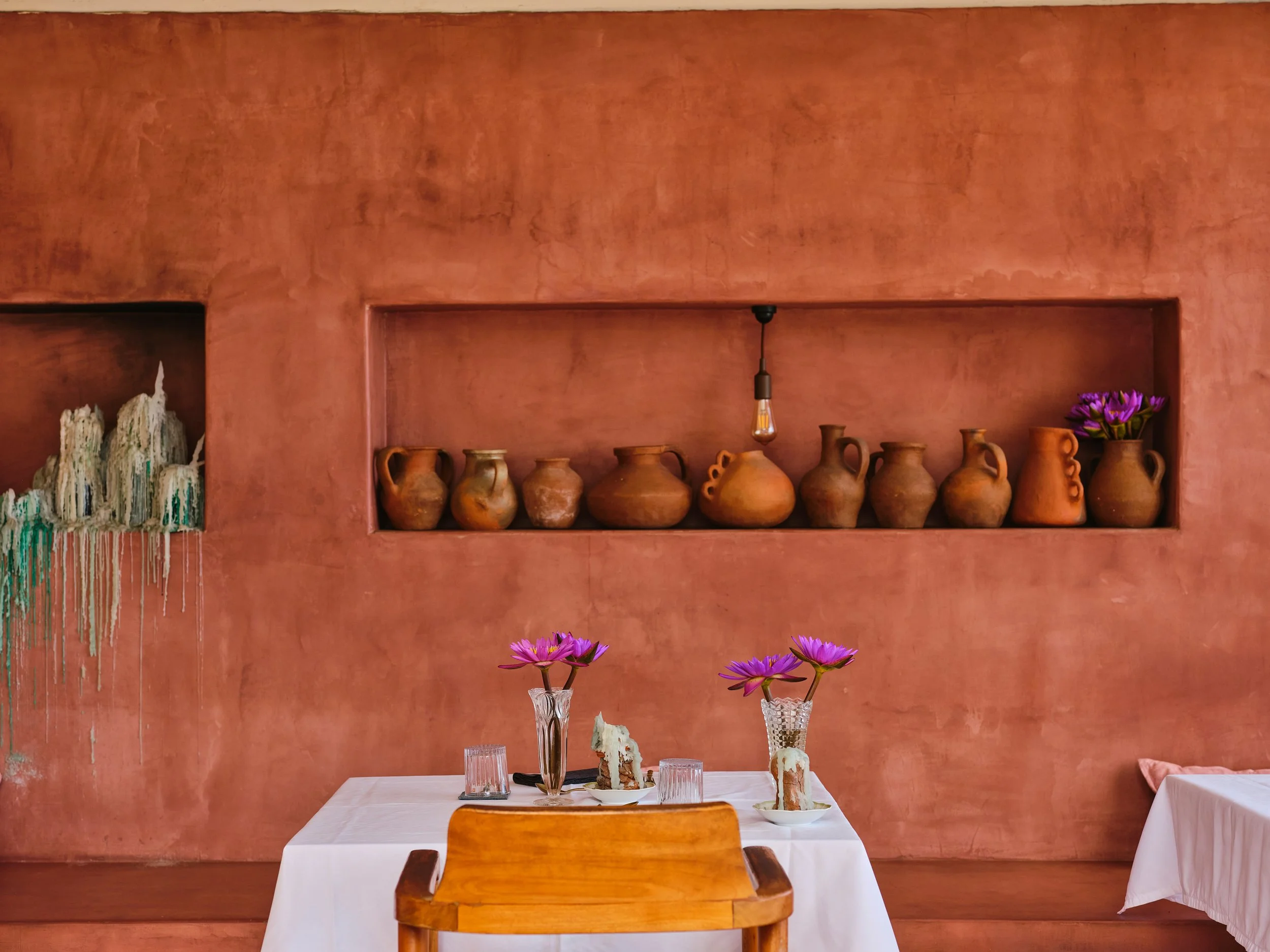 A restaurant table with a white tablecloth, two purple flowers in a glass vase, and two glasses. Behind, a pink textured wall with a built-in shelf holding terracotta pots and purple flowers, and a single hanging light bulb.