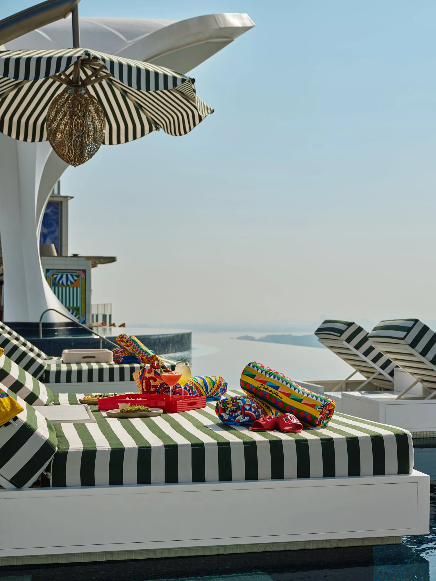 Poolside lounge area with striped cushions, umbrellas, and colorful towels, overlooking a body of water on a sunny day.