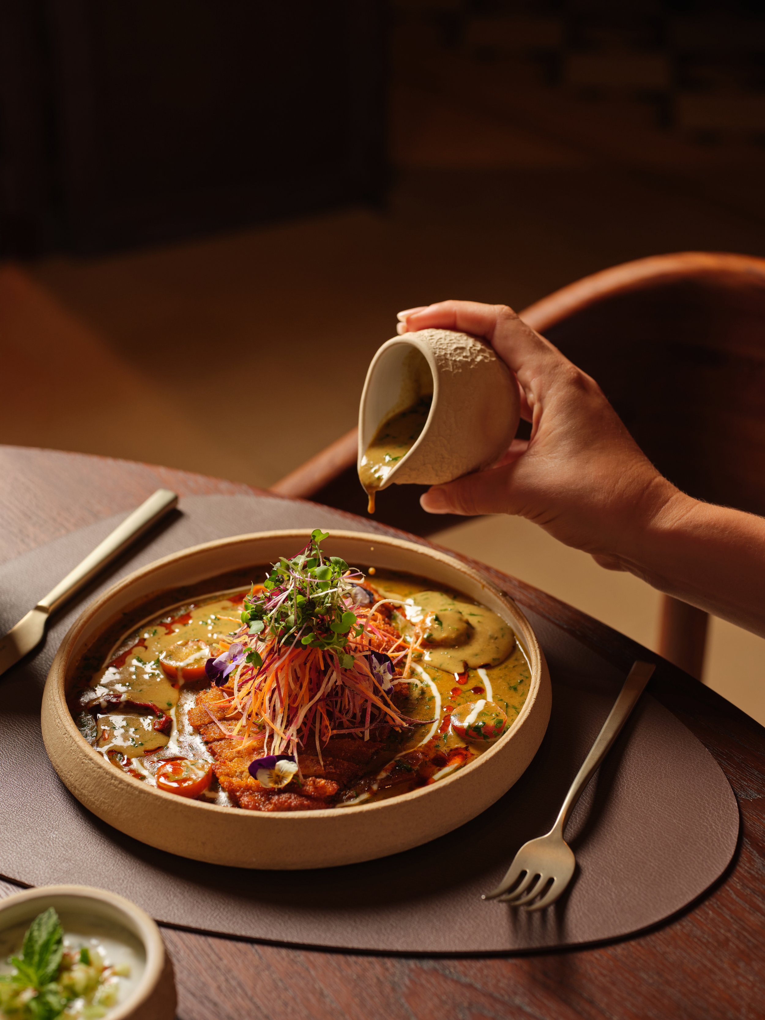 A person pouring green sauce over an Asian-inspired dish with vegetables, microgreens, and edible flowers.