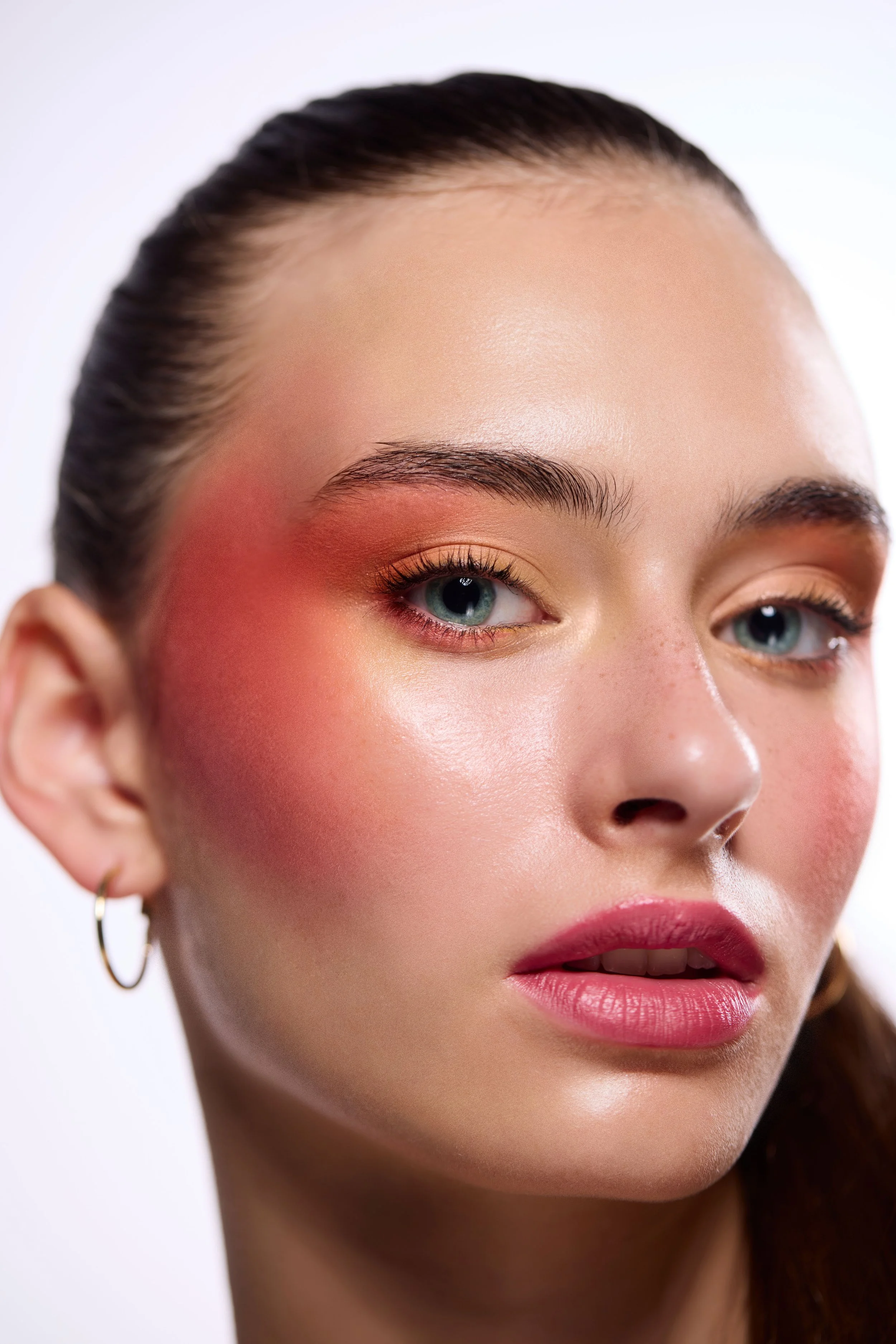 Close-up of a young woman with blue eyes, styled hair, hoop earrings, and natural makeup with pink lipstick and eyeshadow.