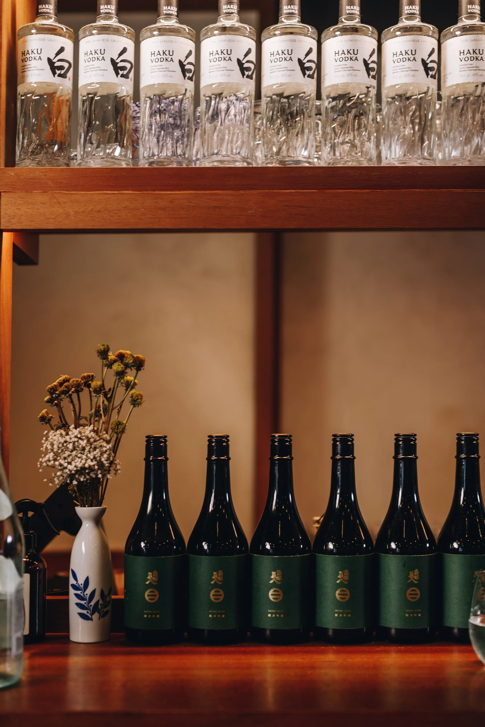 Bottles of Haku vodka on a wooden shelf with a small vase of dried flowers beside them.