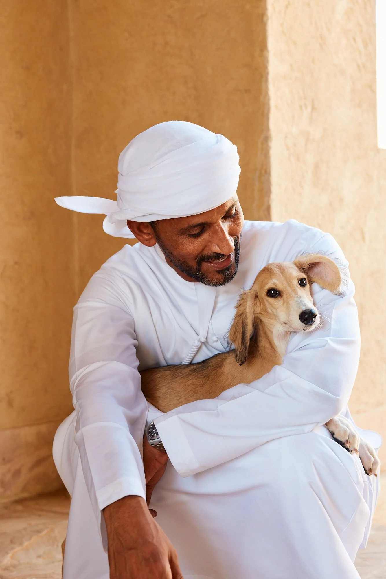 A man dressed in white traditional attire, wearing a white turban, sitting on the floor and holding a small tan and white dog in his lap, with warm beige walls in the background.