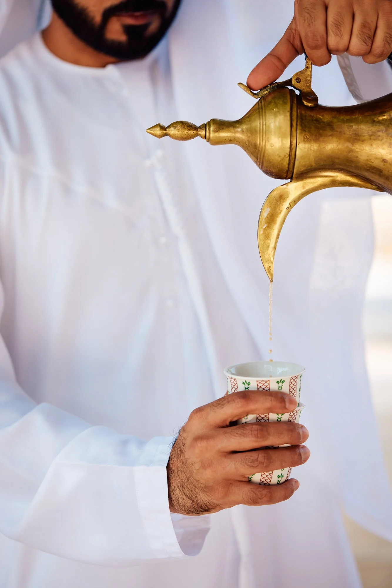 A man in traditional white Middle Eastern attire is pouring a beverage from a brass coffee pot into a decorated paper cup.