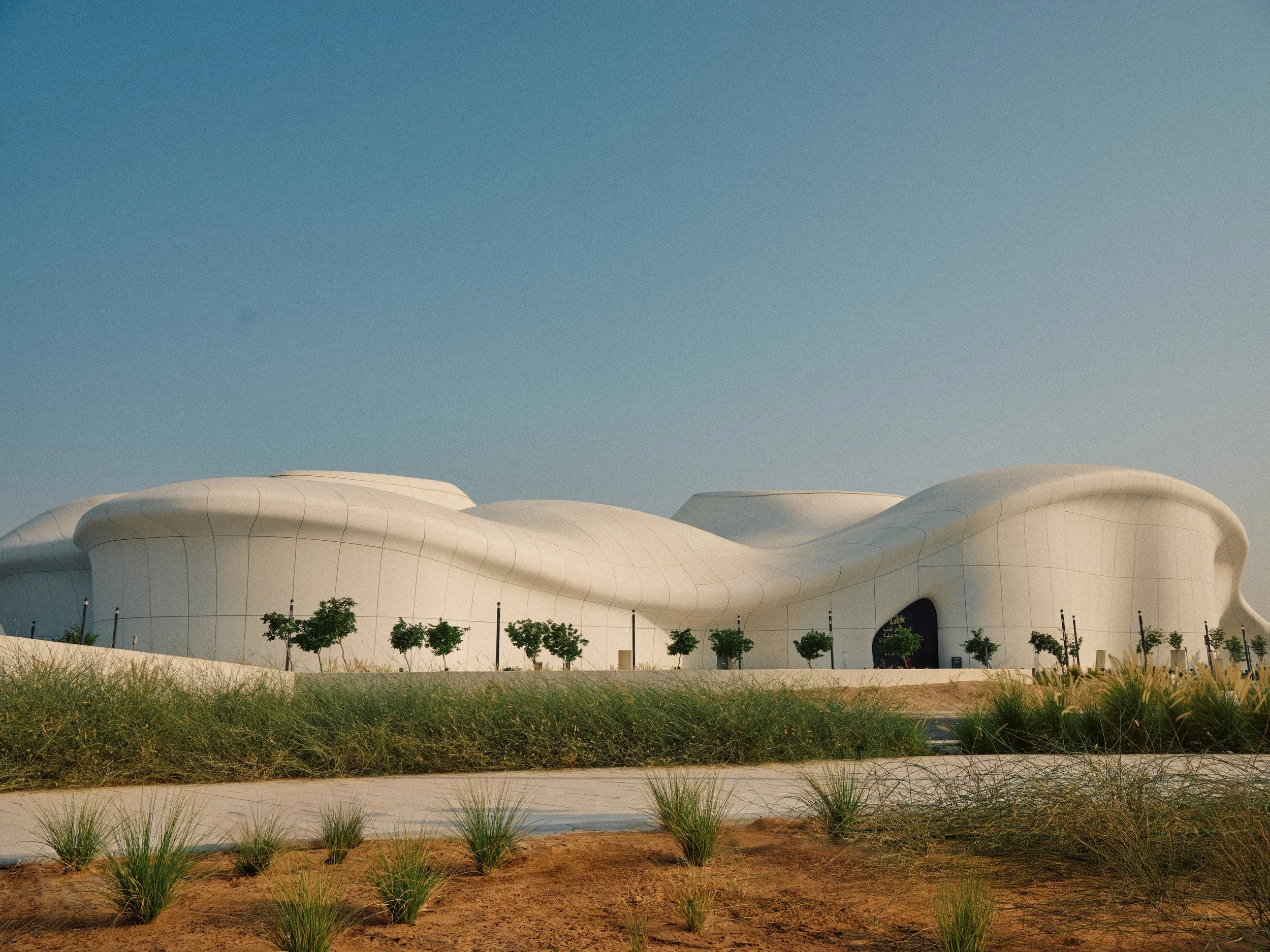 Modern, white, curvaceous building with green trees lining its exterior, set against a clear sky.