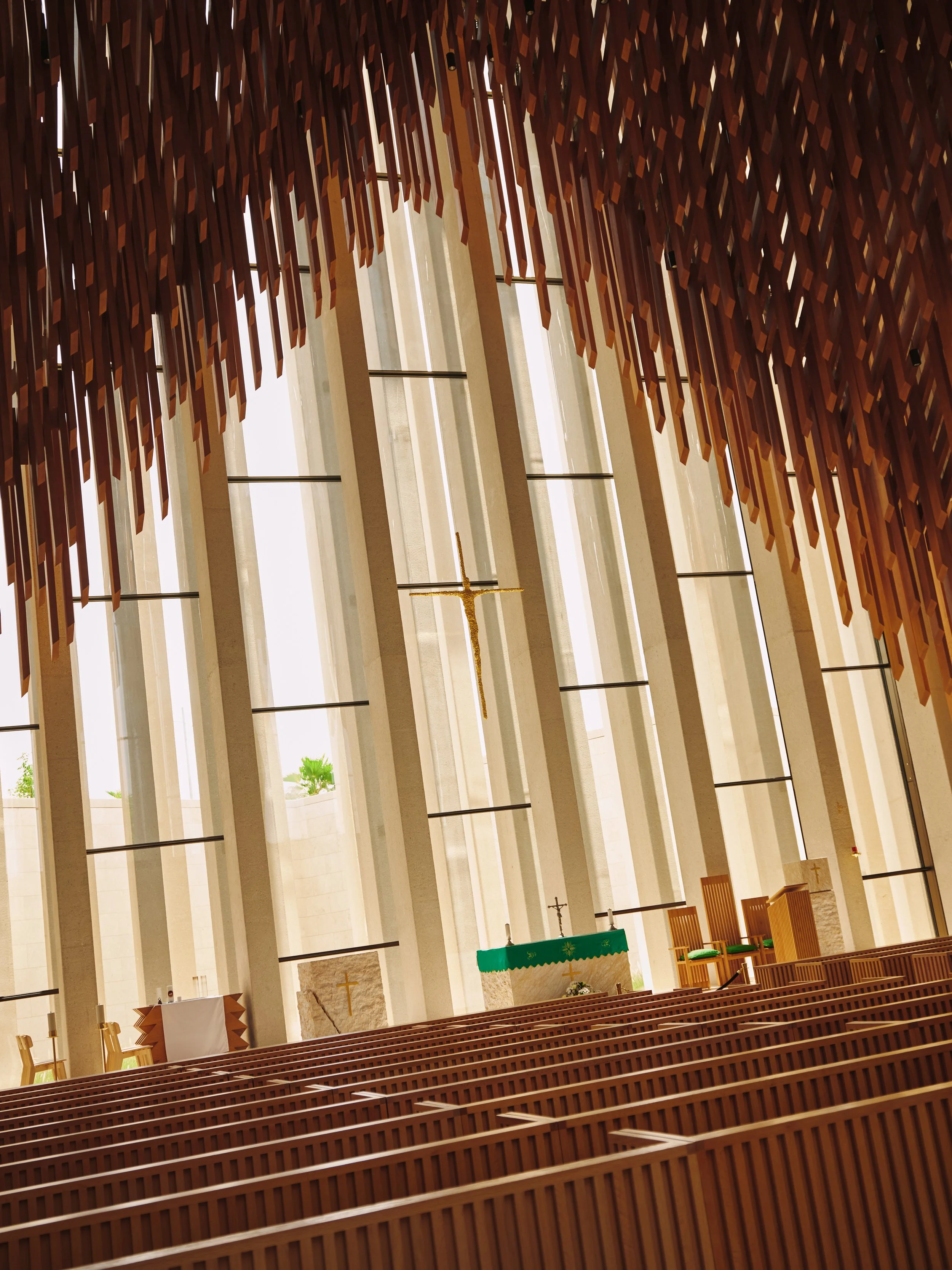 Interior of a modern church with wooden pews, tall vertical windows, and an altar with a green cloth, a cross, and a hanging crucifix, under a ceiling made of wooden slats.