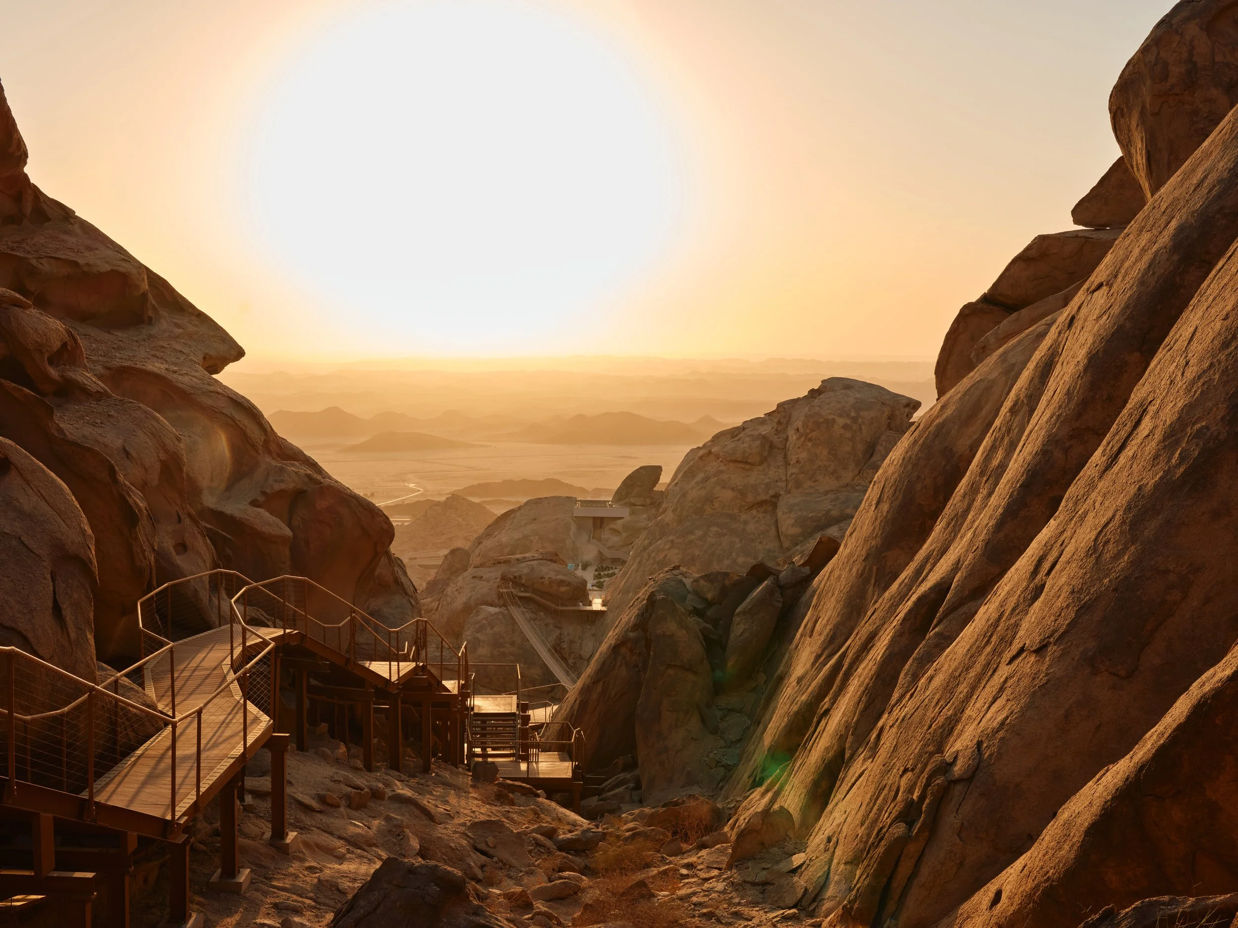 Rocky desert landscape with a pathway and stairs, under a setting sun, with distant mountains and a clear sky.