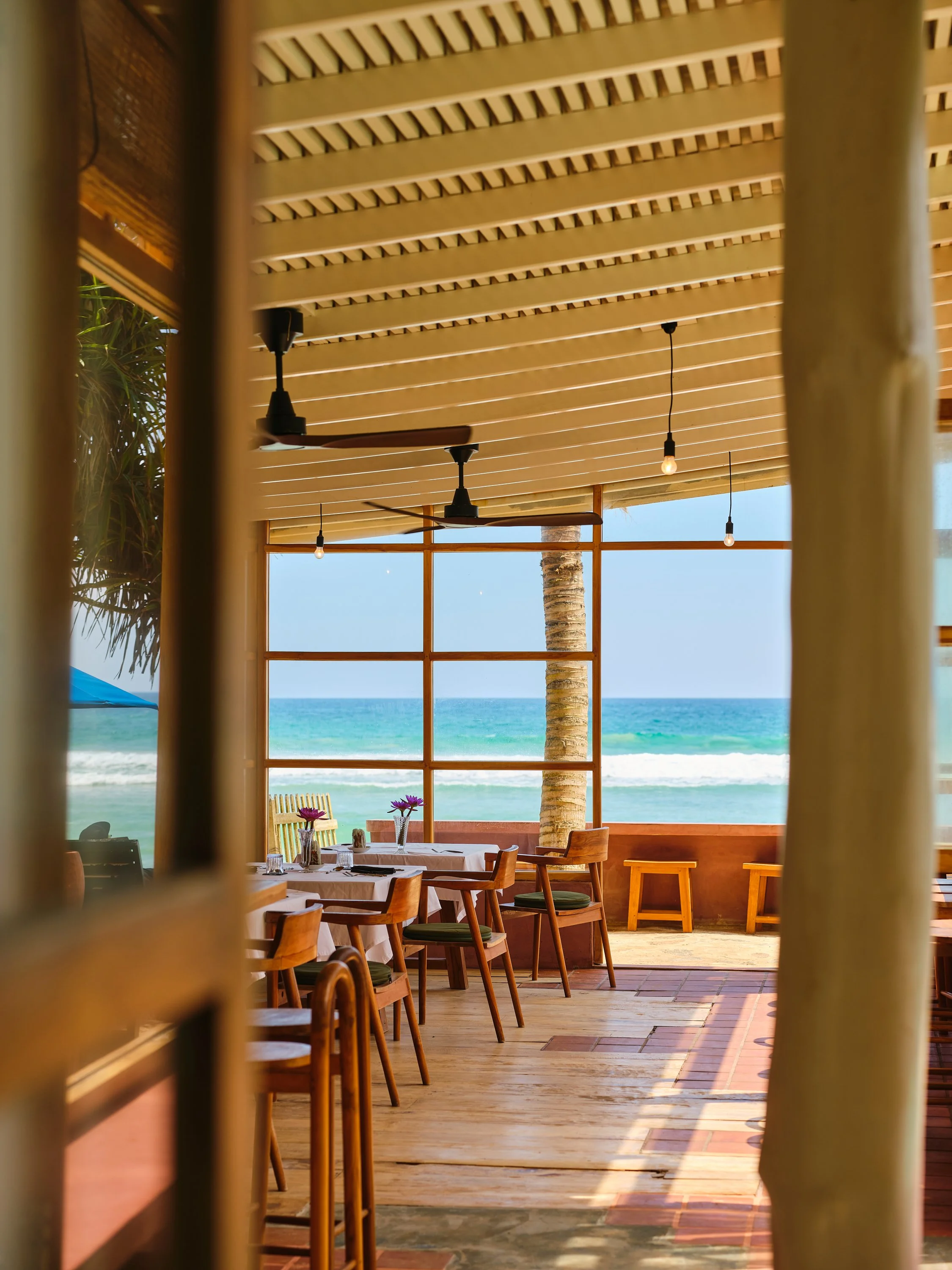 Interior of a beachside restaurant with wooden furniture, overlooking the ocean through large windows, decorated with flowers, ceiling fans, and hanging light bulbs.