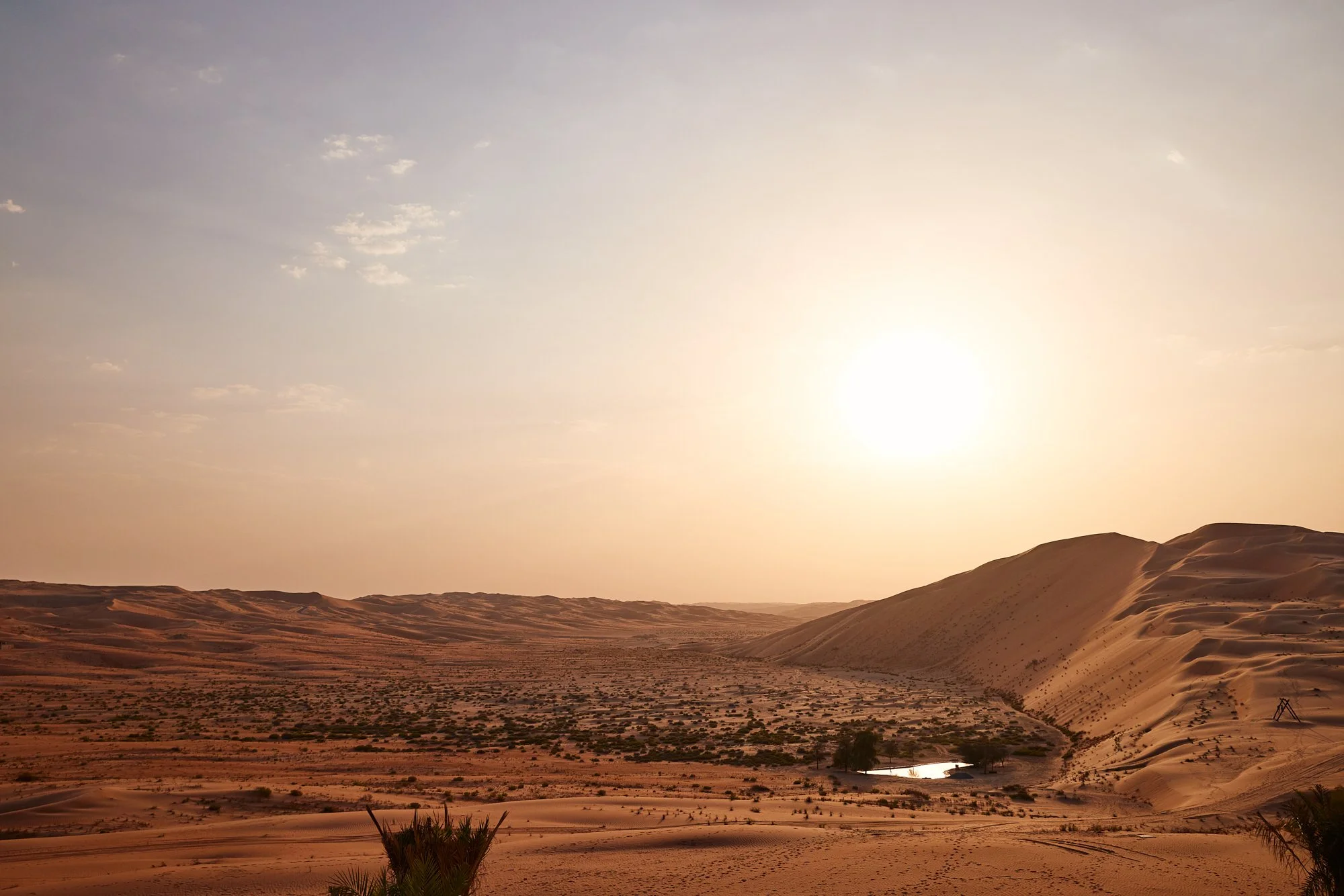 A desert landscape at sunset with sandy dunes, sparse vegetation, and a small water pond reflecting the sky, under a bright sun in a mostly clear sky.