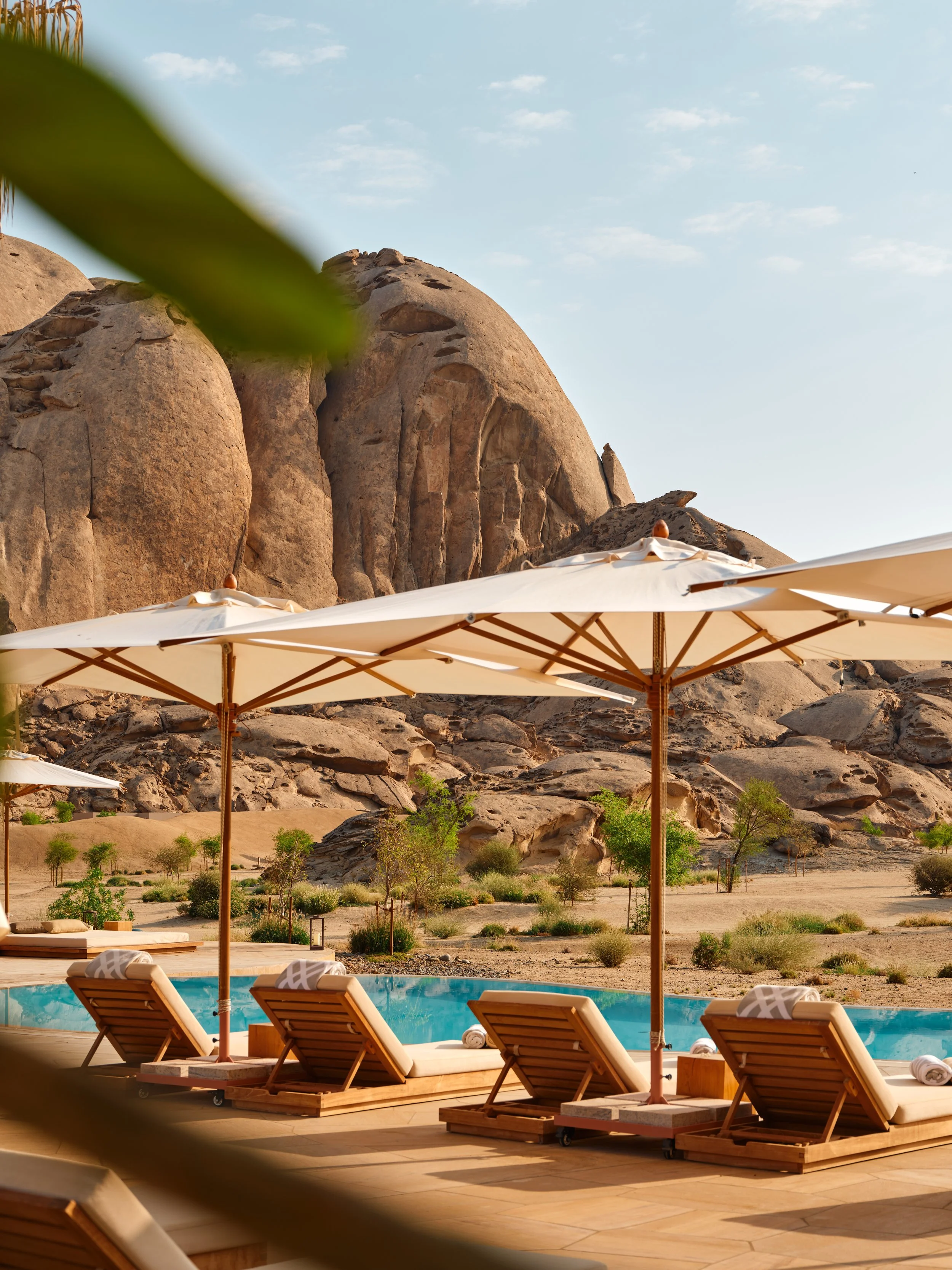 Poolside area with lounge chairs and umbrellas, desert landscape with rocks and sparse trees, mountains in the background and a partly cloudy sky.