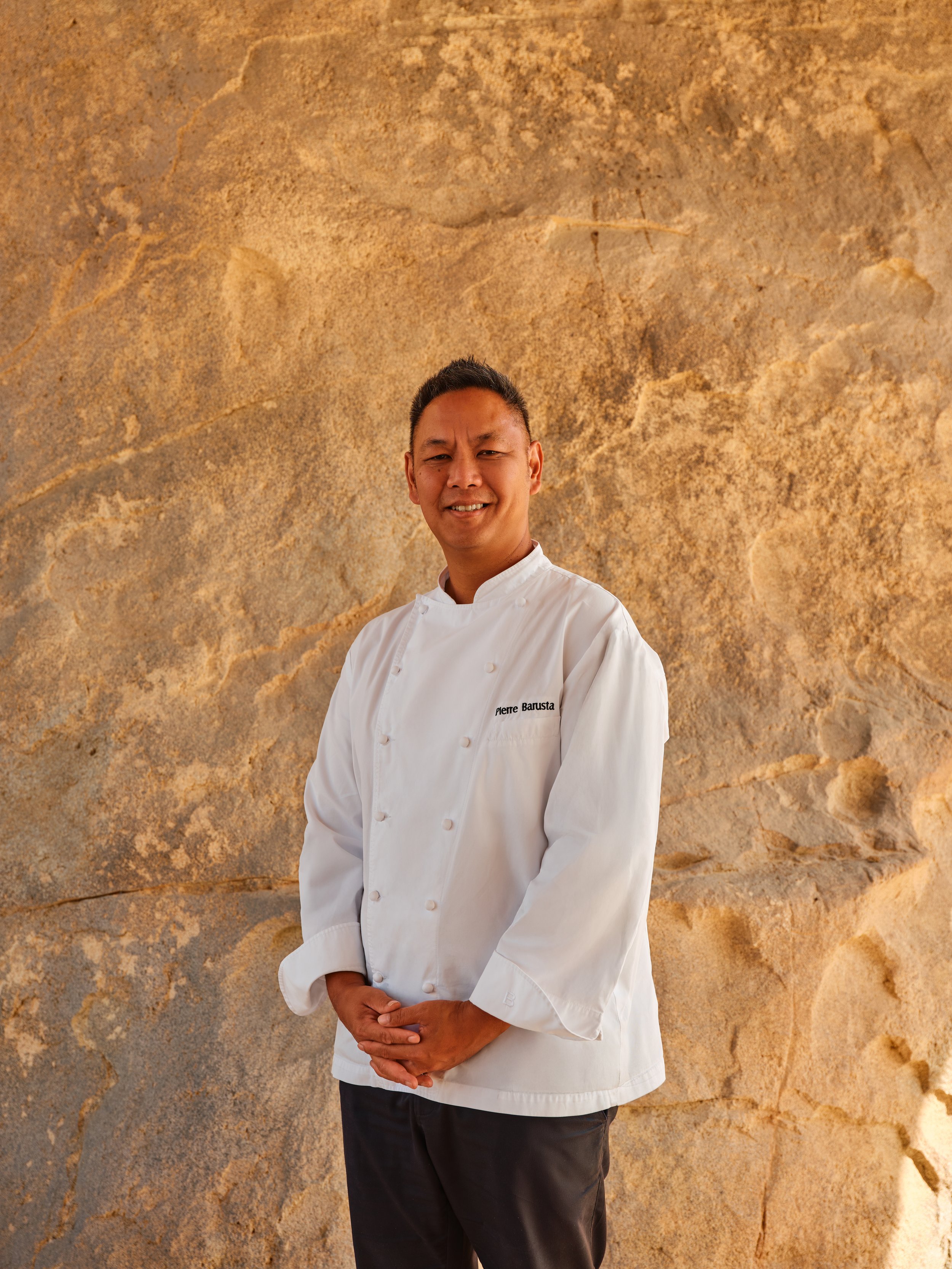 A chef in white uniform standing in front of a textured beige stone wall.