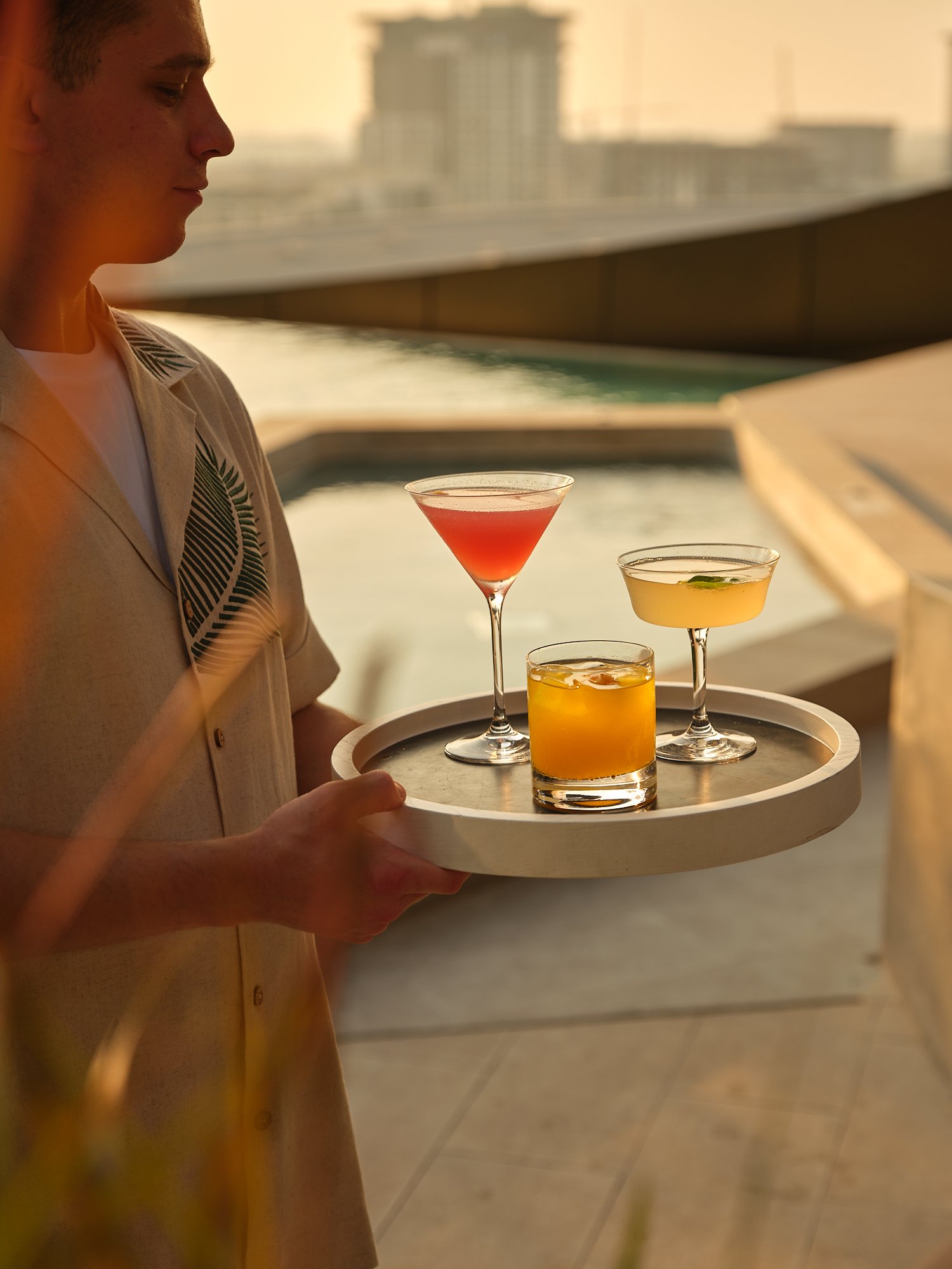 A person holding a tray with four colorful cocktails near a pool or body of water during sunset.
