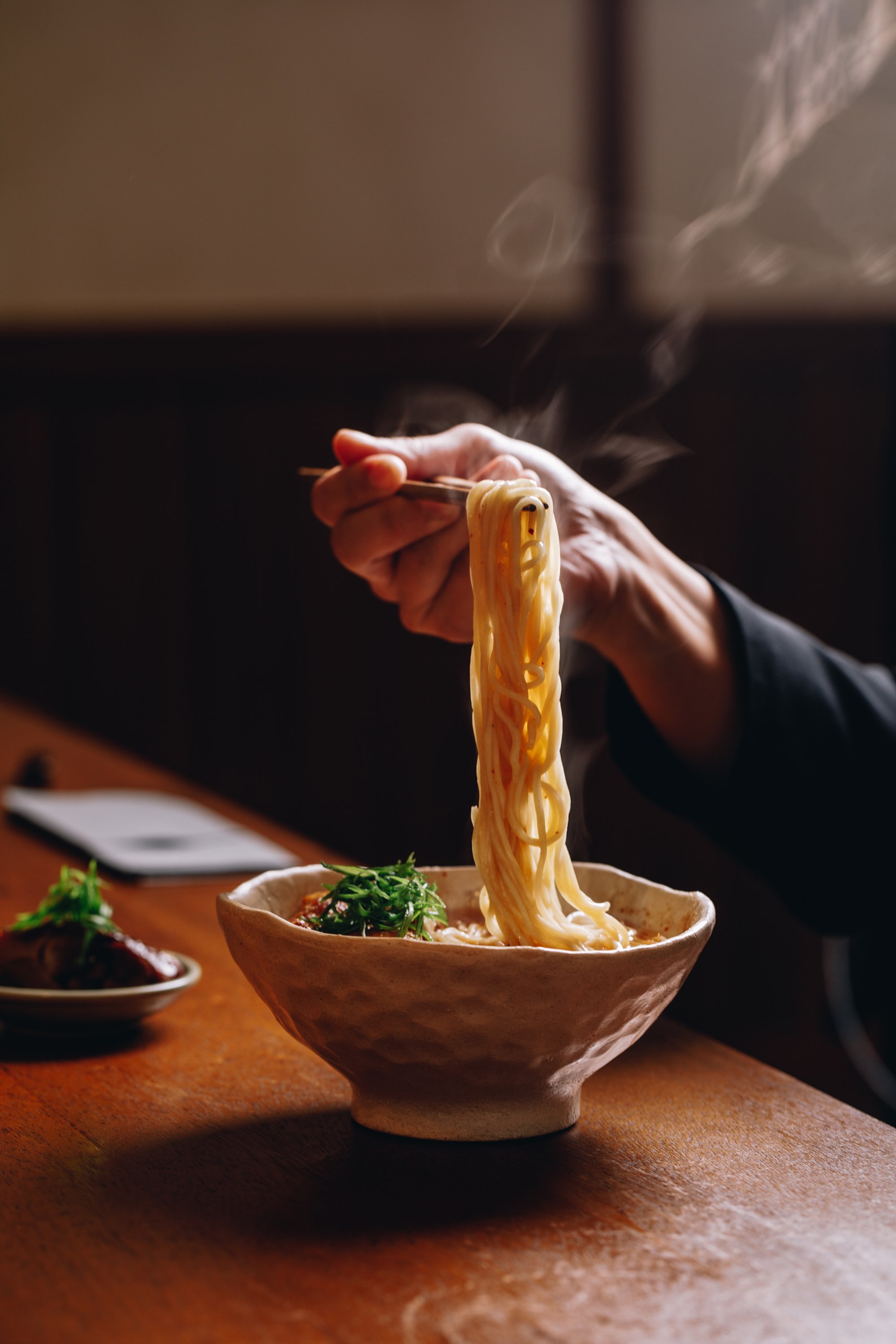 A hand holding chopsticks lifting noodles from a bowl of ramen, with steam rising. There is a small plate of meat garnished with herbs on the side and a smartphone on a wooden table.