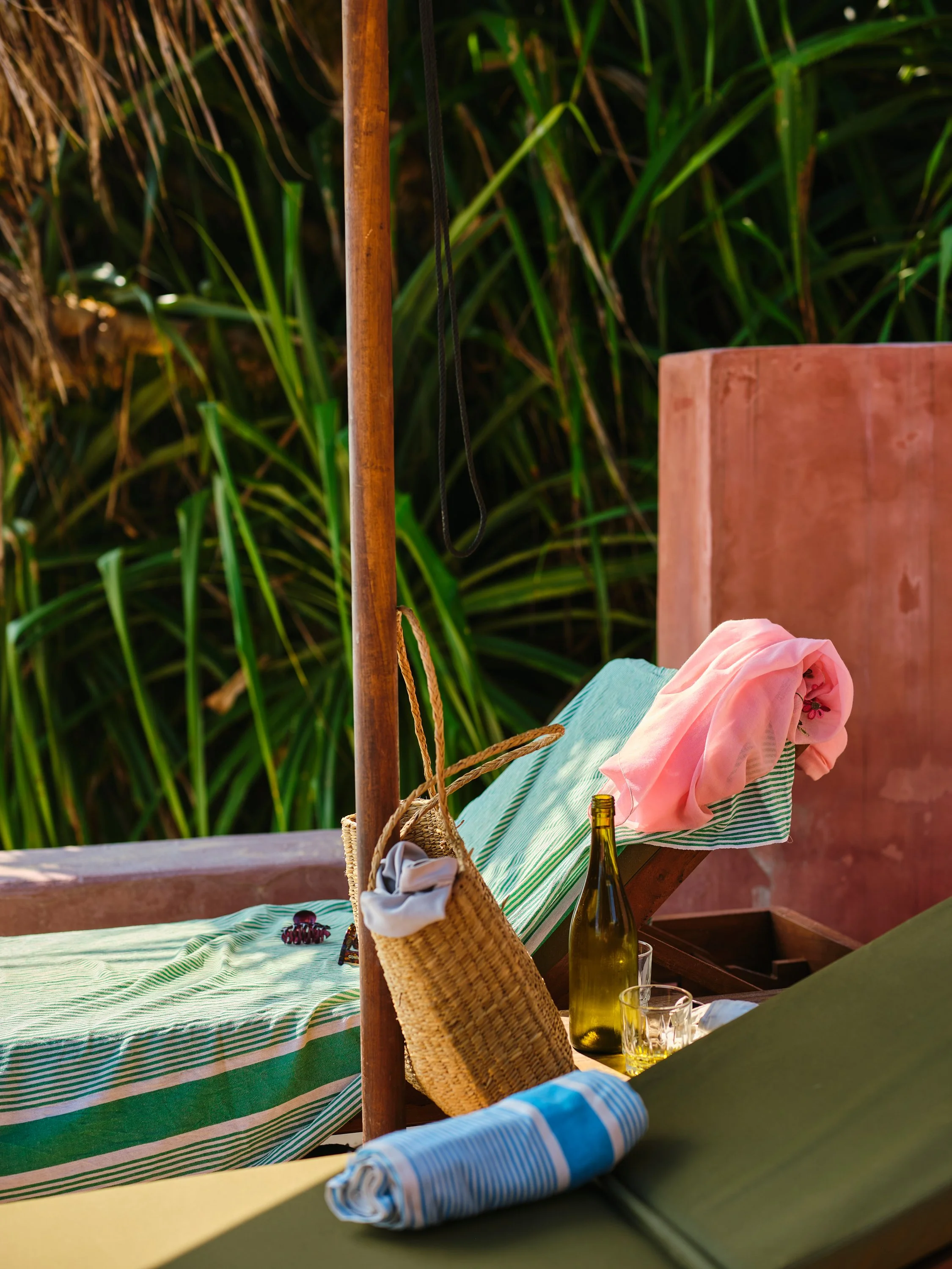 A lounge chair with a pink cloth, a glass bottle, and a glass on a table, surrounded by greenery.