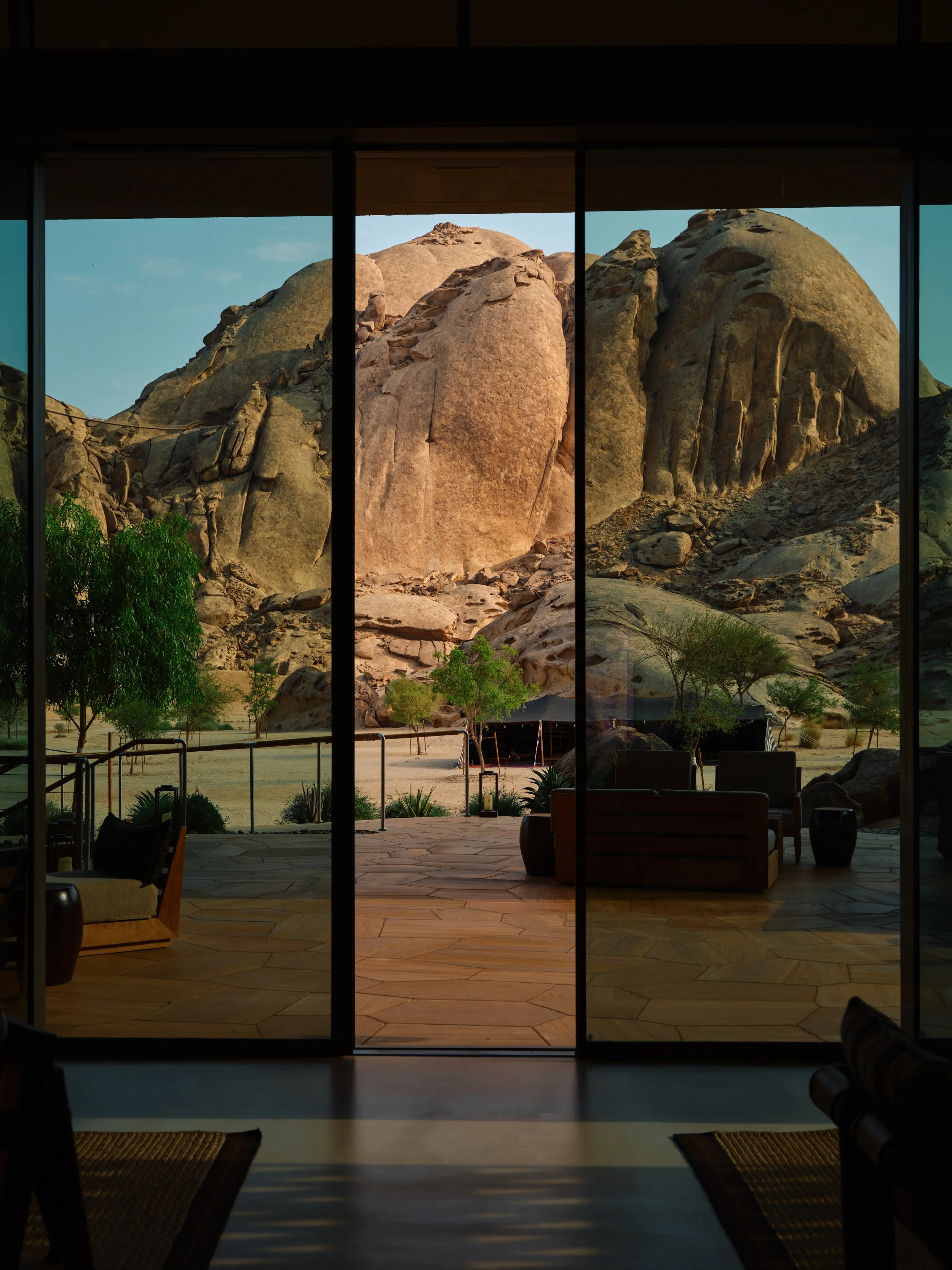 View of desert landscape with large rock formations and sparse trees, seen through glass sliding doors from inside a room with wooden flooring and furniture.