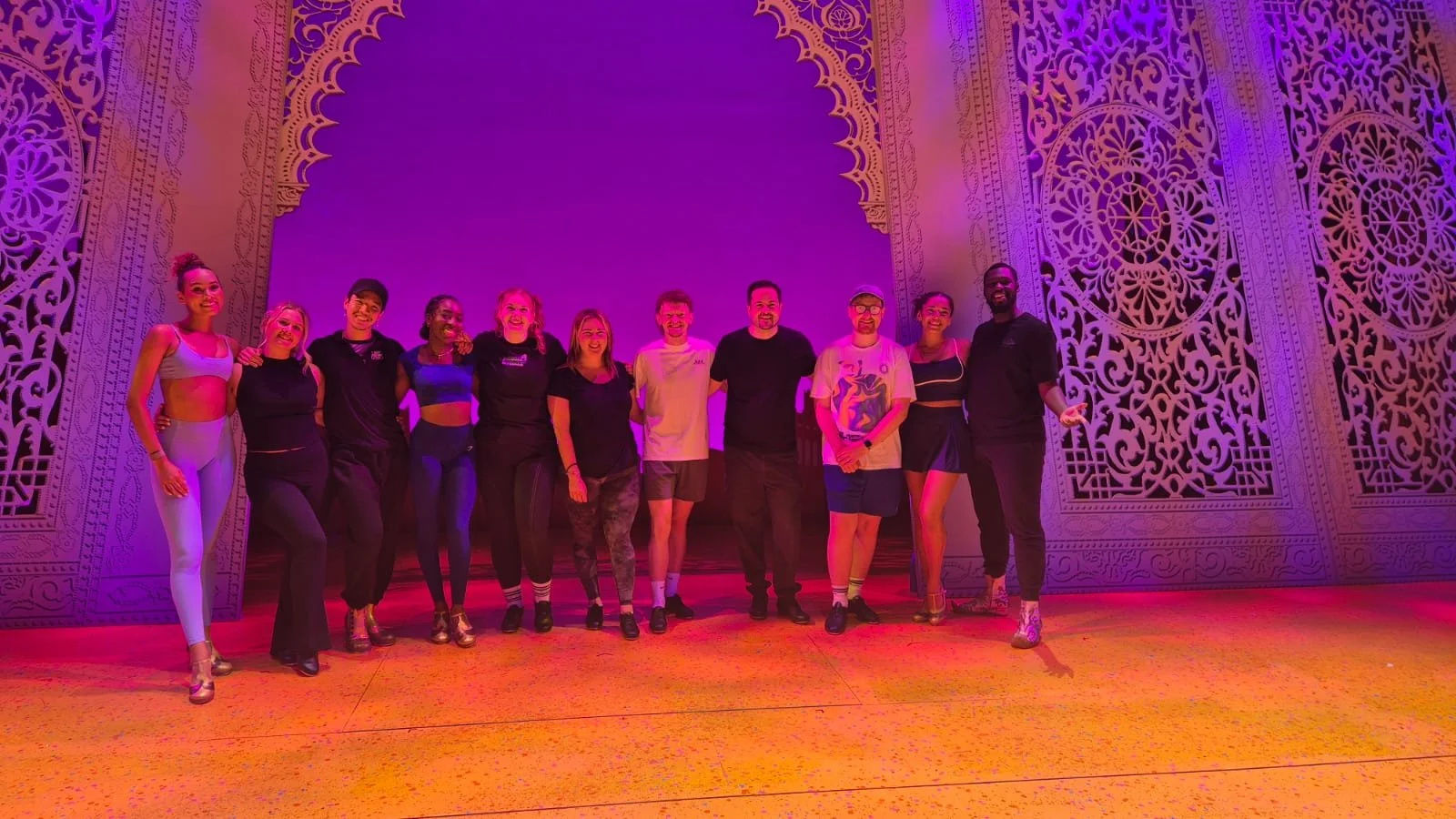 Group of people standing on stage with colorful lighting and ornate wall designs in the background.