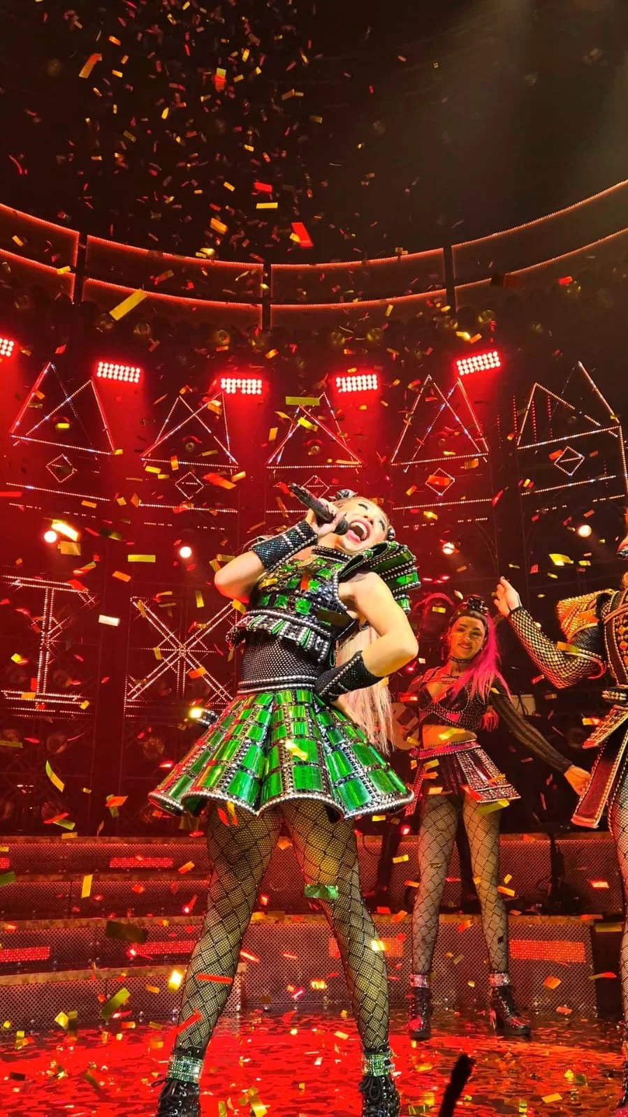 A performer singing on stage with colorful costumes, surrounded by red lights, confetti, and fellow performers in similar attire, under a geometric light display.