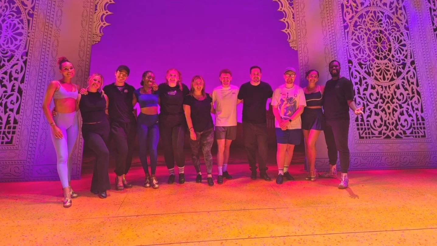 Group of twelve people standing together on a stage, smiling and posing for a photo with a decorative backdrop.