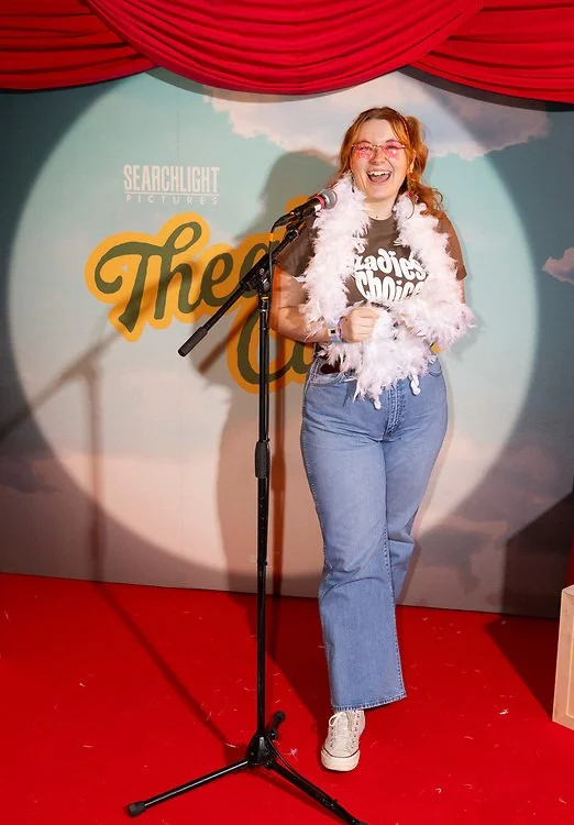 Woman with red hair, glasses, and a white feather boa, smiling and standing on stage with a microphone, in front of a backdrop with writing and red curtains.