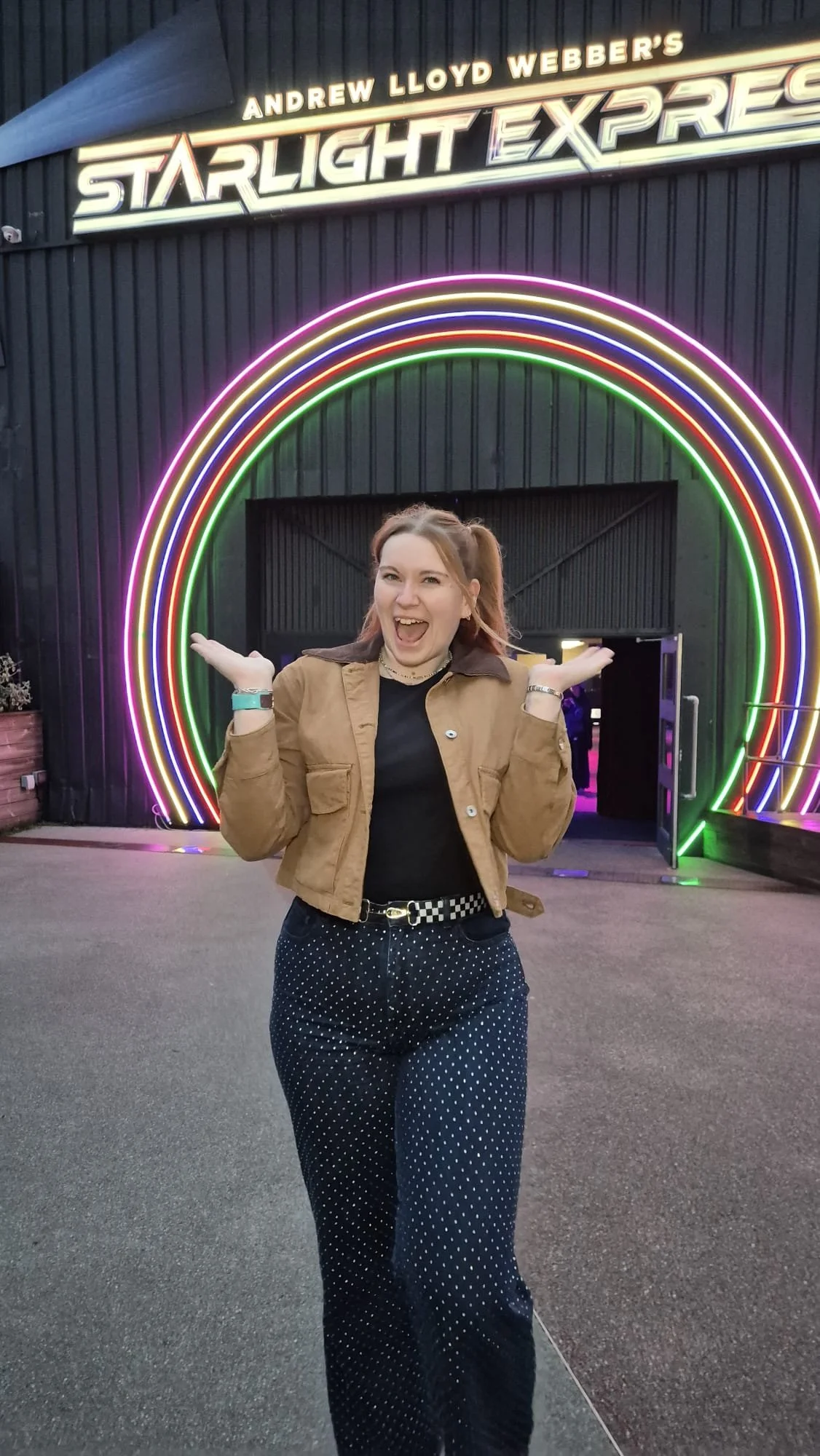 A woman smiling and posing with arms raised in front of a neon-lit rainbow arch outside 'Starlight Express' theater, with a sign reading 'Andrew Lloyd Webber's' above.