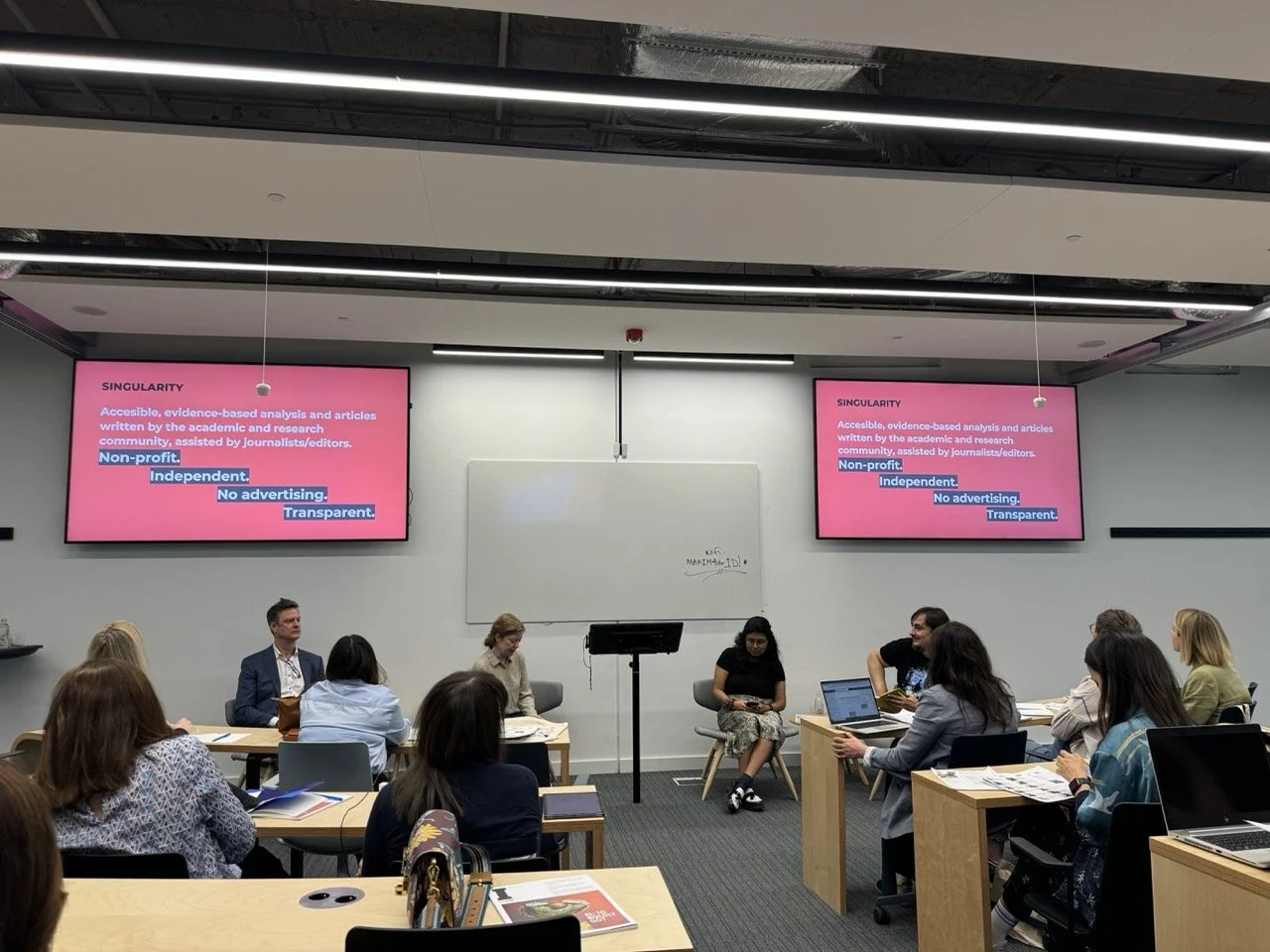 A classroom or seminar room with multiple people seated at tables, facing a whiteboard and two large screens displaying a pink slide with text about singularity and non-profit organizations.