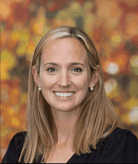 A smiling woman with long blonde hair, wearing pearl earrings, in front of a colorful blurred autumn background.
