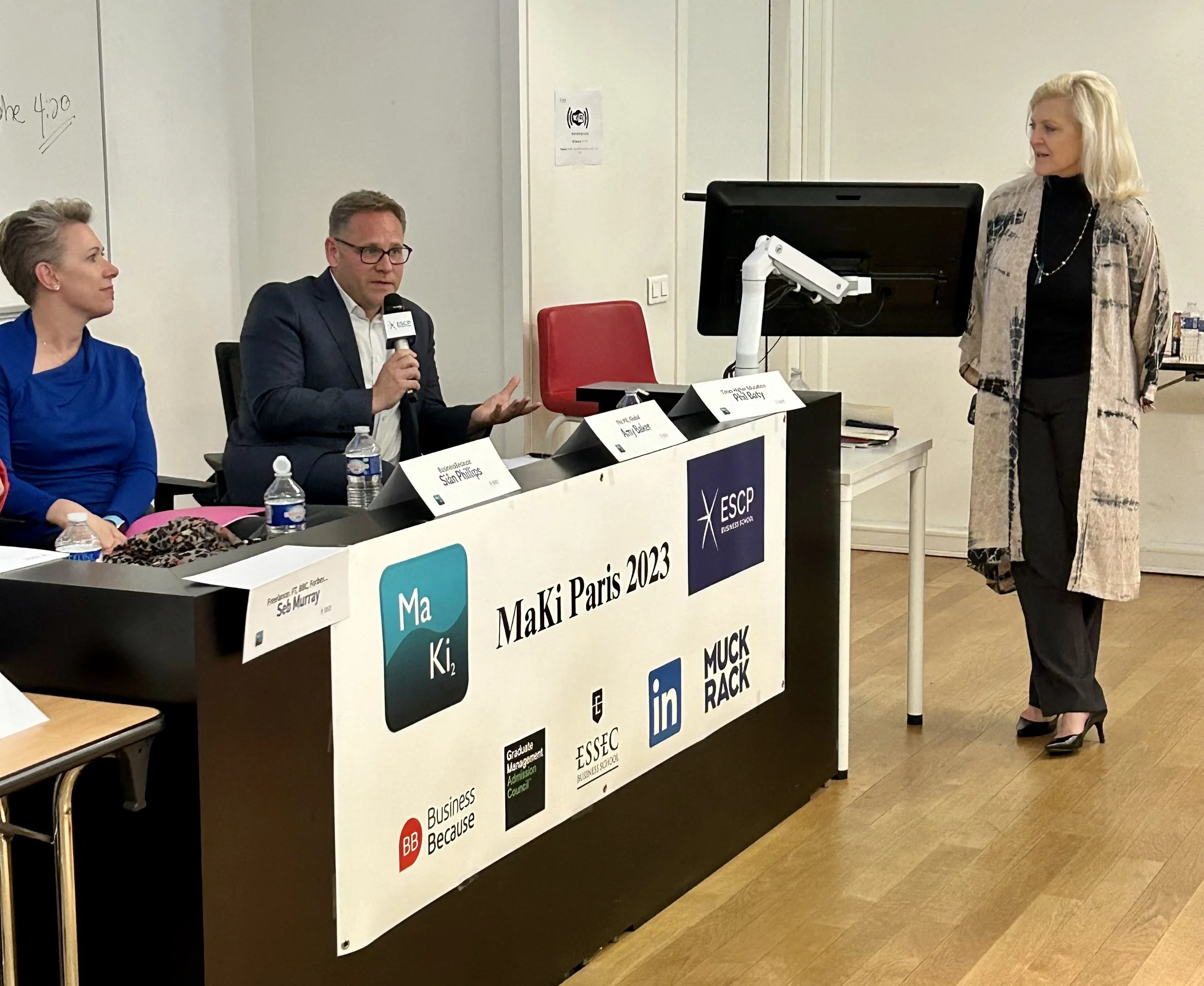 A panel discussion at Maki Paris 2023 event with three women and one man seated at a table, one woman standing next to a monitor, and a banner displaying event information and logos.