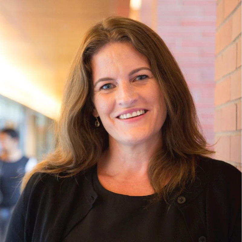 A woman with shoulder-length brown hair, smiling, wearing a black top and earrings, standing indoors near a brick wall with a blurred background.