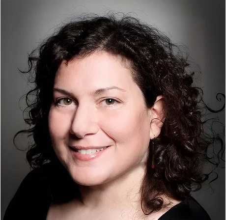 Portrait of a woman with curly dark hair smiling against a gray background.
