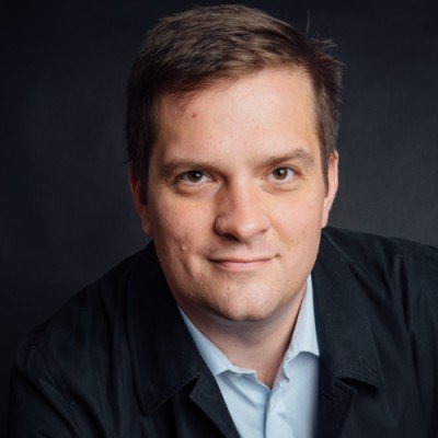 A professional headshot of a man with short brown hair, light skin, wearing a light blue collared shirt and a black blazer, against a dark background.