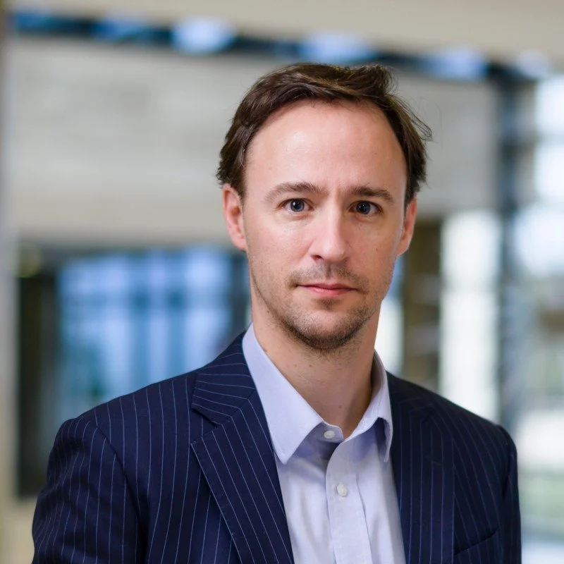 A professional man in a pinstripe suit poses for a portrait in an indoor office setting.