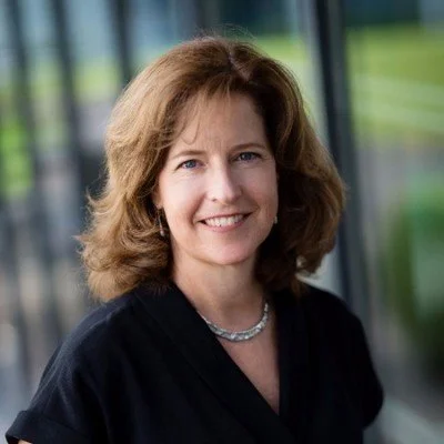 A woman with shoulder-length brown hair, smiling, wearing a black top and a silver necklace, standing indoors near large windows