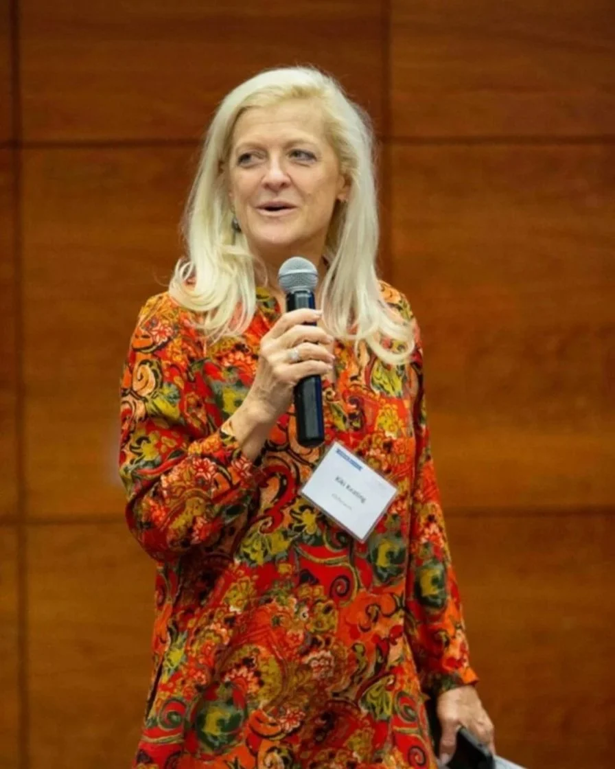 A woman with long blonde hair wearing a colorful, patterned dress, holding a microphone and speaking at an event.