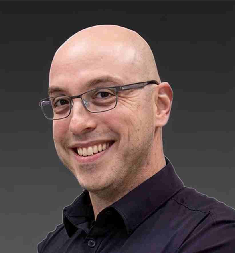 A smiling man with glasses and no hair, wearing a black shirt, against a dark background.