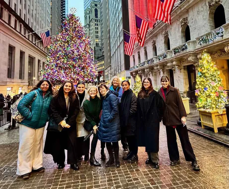 Group of nine women standing outdoors in front of decorated Christmas trees and a large, colorful Christmas tree in a city street with tall buildings and American flags displayed.
