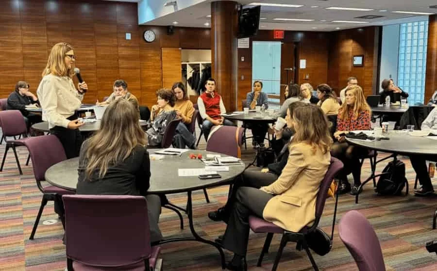 A woman holding a microphone and speaking to a seated audience in a conference room with wooden walls and round tables surrounded by chairs.