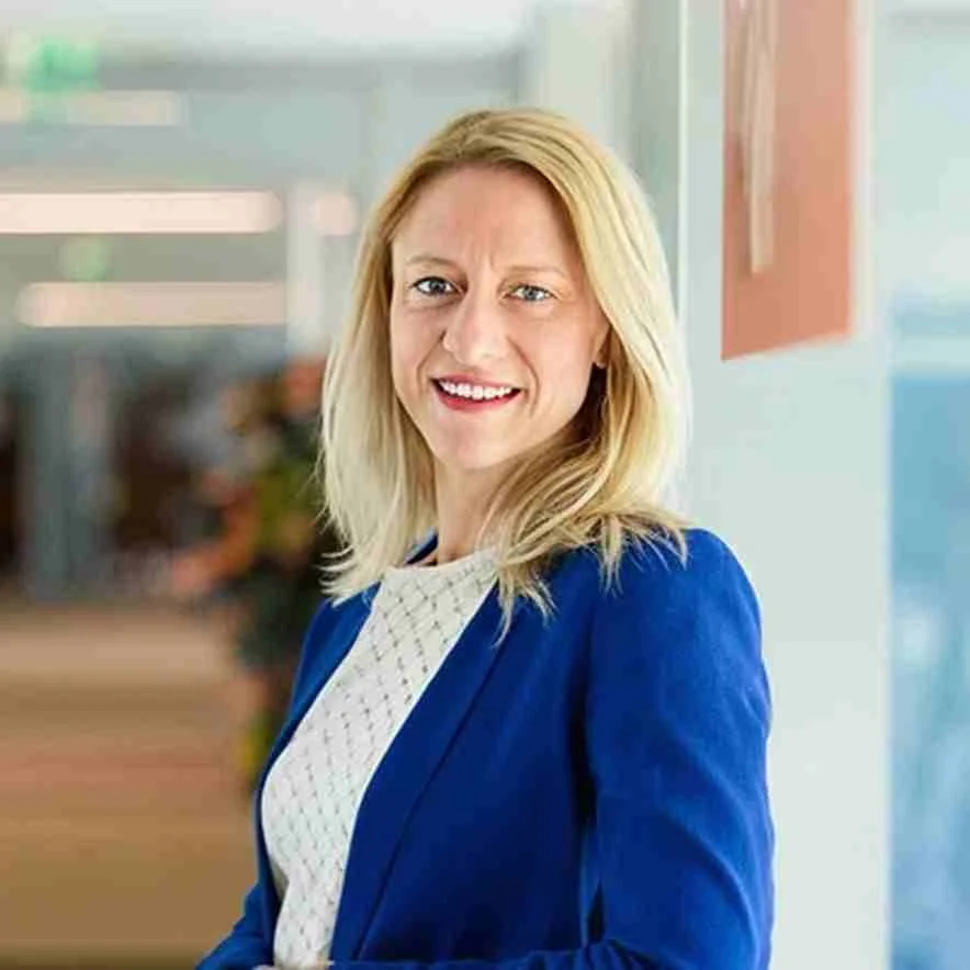 A smiling woman with blonde hair, wearing a blue blazer and white blouse, standing in a bright indoor space.