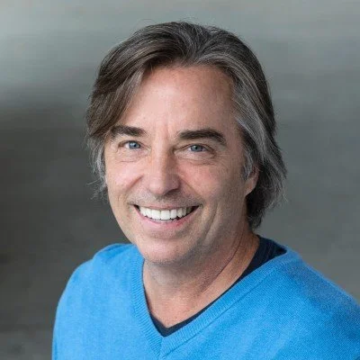 Headshot of a middle-aged man with gray hair, blue eyes, and a broad smile, wearing a blue shirt.