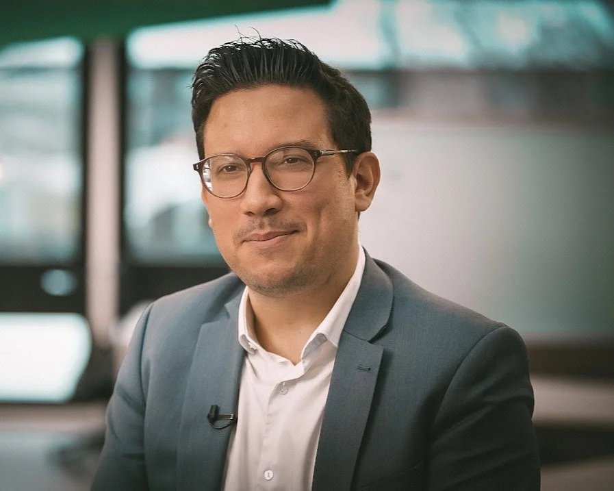 A man with dark hair, glasses, and light skin wearing a gray suit and white shirt, sitting indoors with blurred windows in the background.